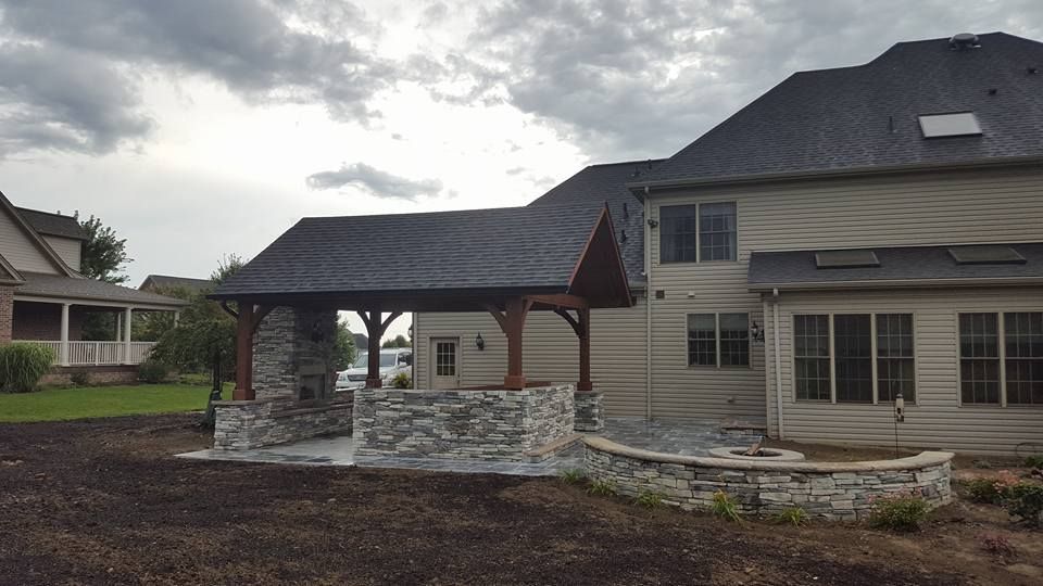 Backyard patio with stone fireplace, attached to a house with a dark roof.