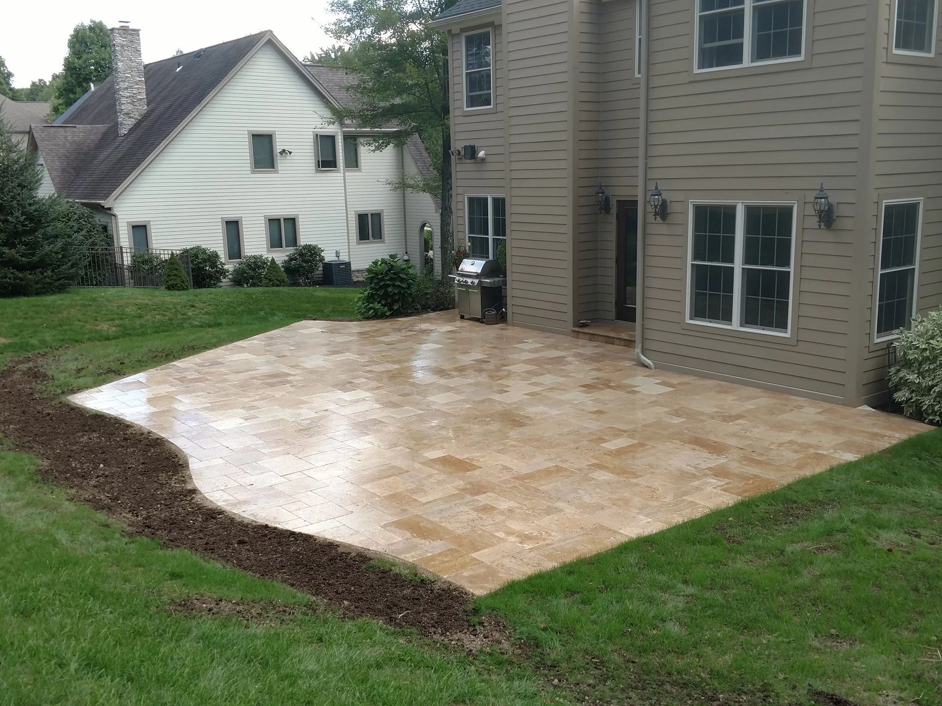 Beige tiled patio next to a tan house with a grill, grass and landscaping, another house in the background.