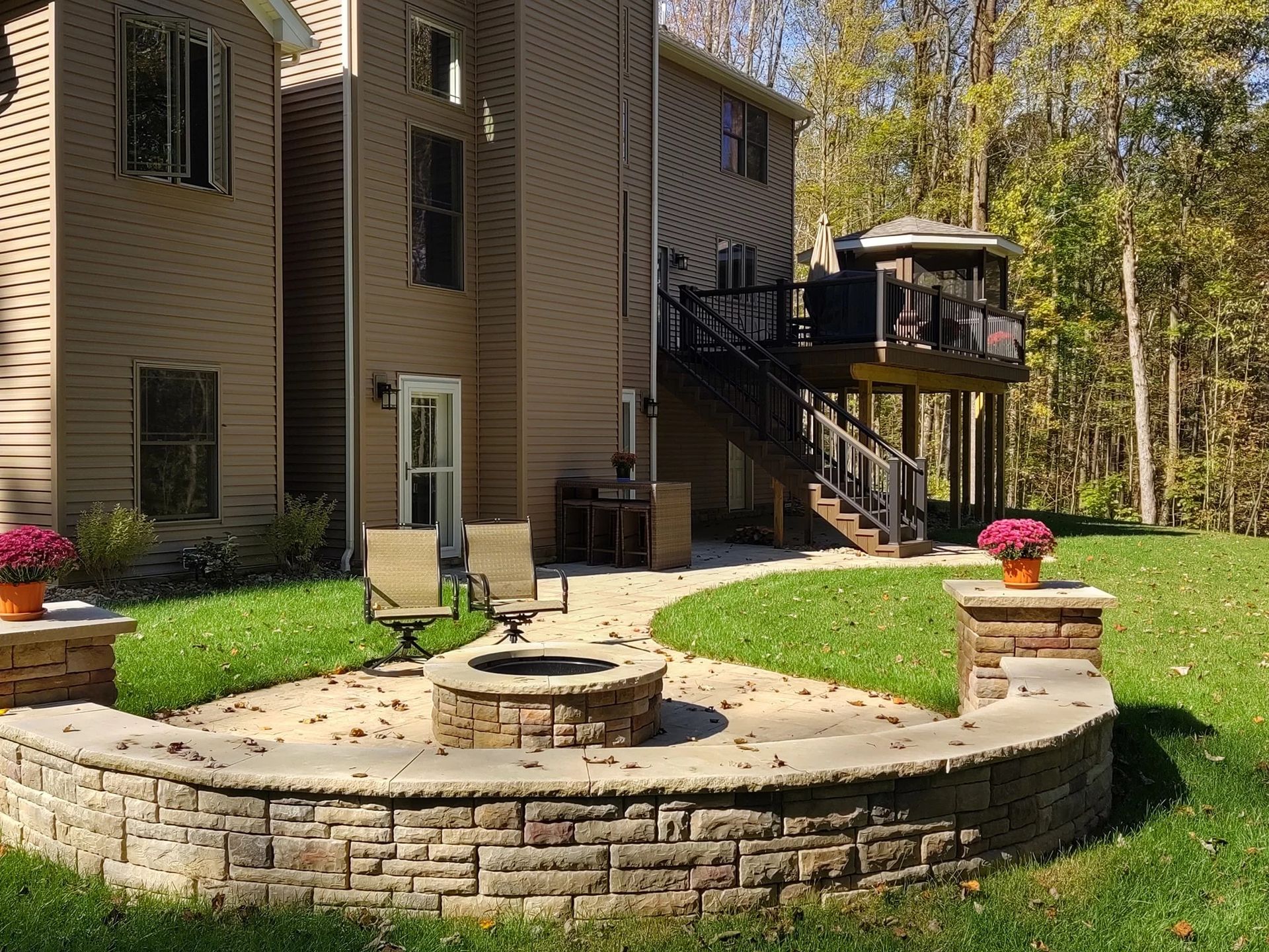 Backyard patio with fire pit, seating, and raised deck with gazebo, surrounded by grass and trees.