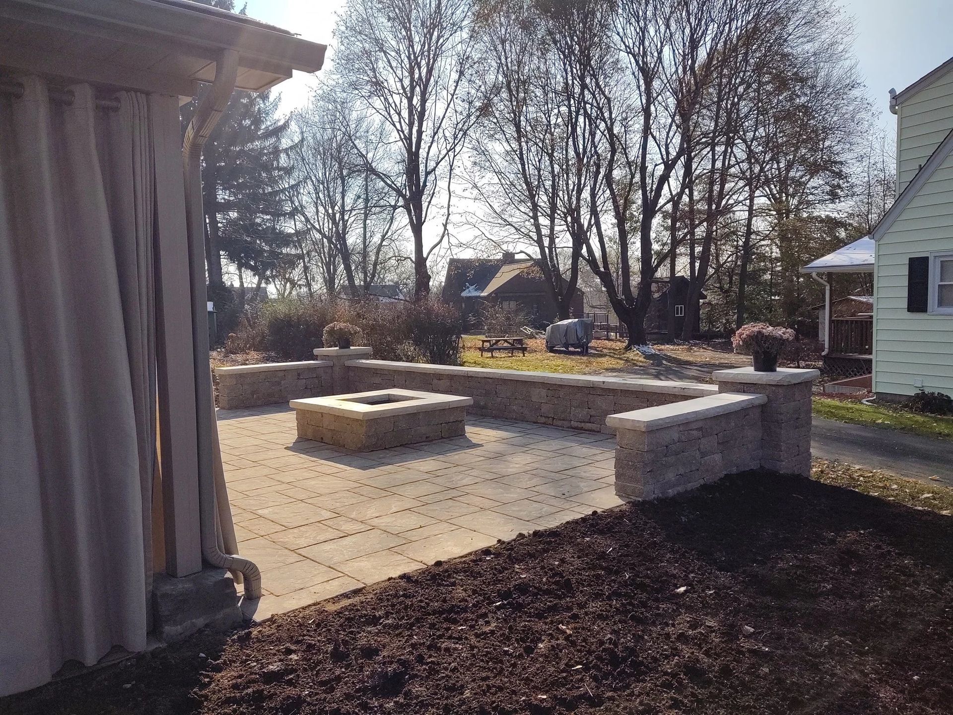 Patio with stone pavers, fire pit, and retaining walls, with a beige curtain and landscaping.