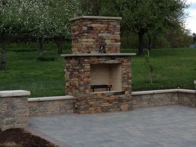 Stone fireplace on patio with retaining wall, green lawn, and trees in the background.