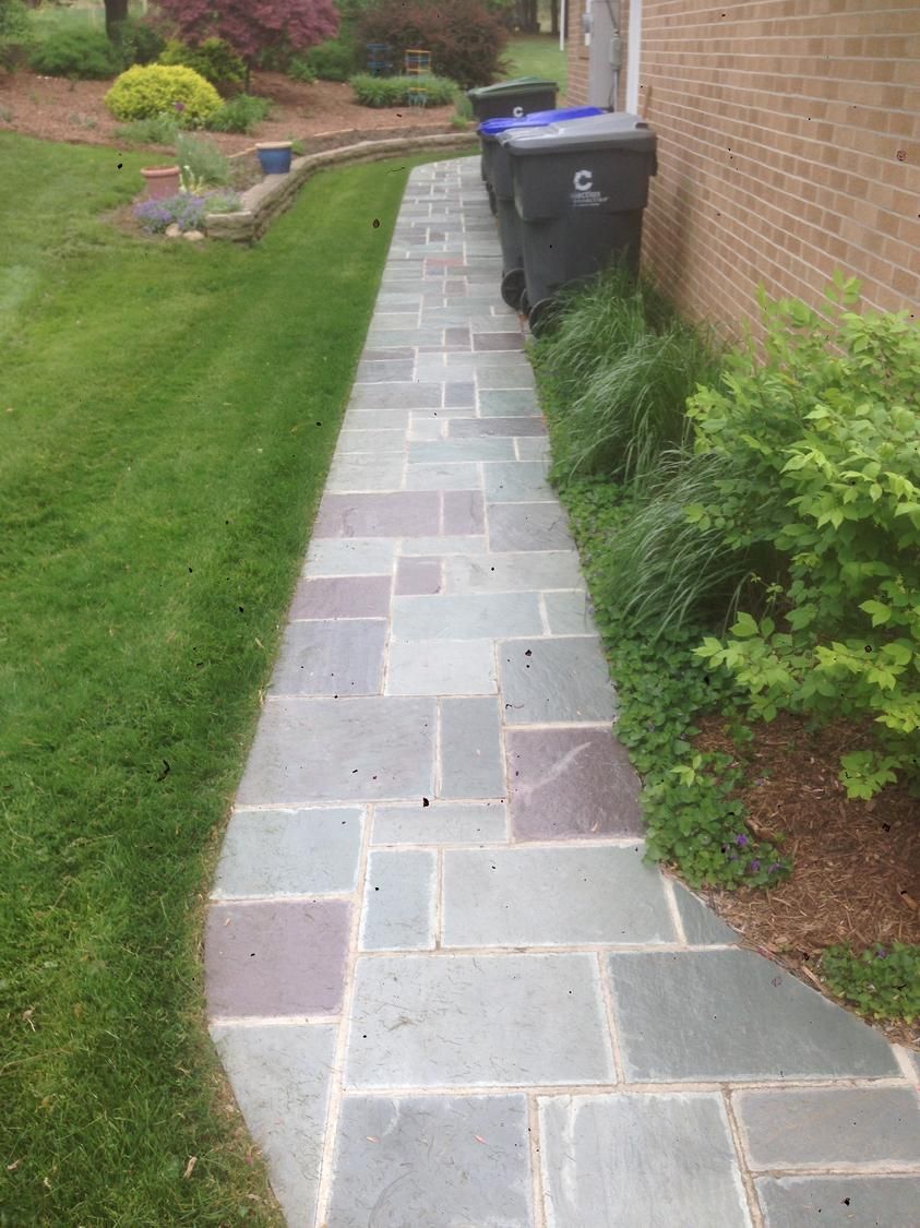 Stone path alongside a brick wall and grass, with trash bins visible at the end.