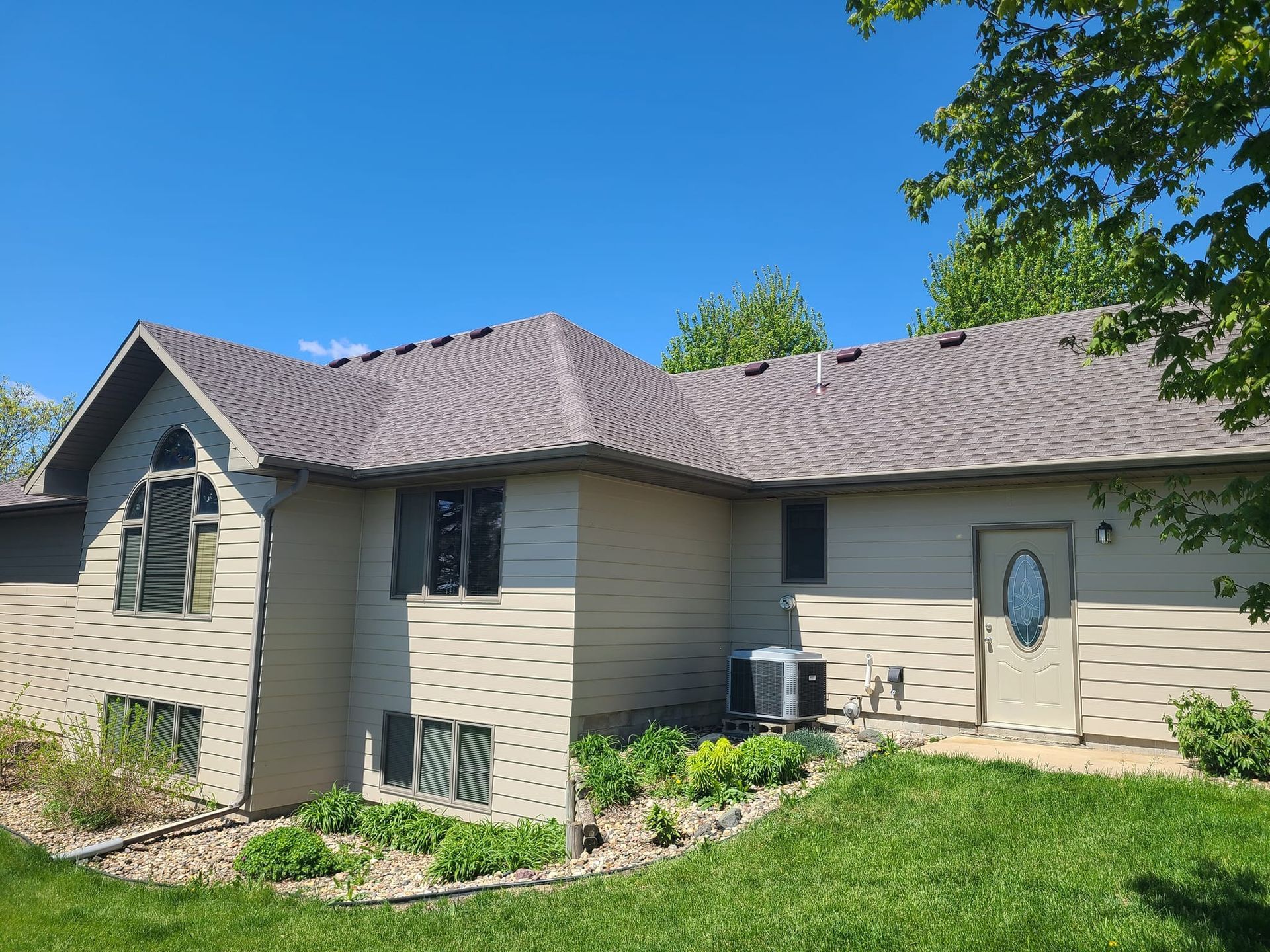 A one-story tan house with a gray shingled roof, a front door, and a landscaped yard under a clear blue sky.