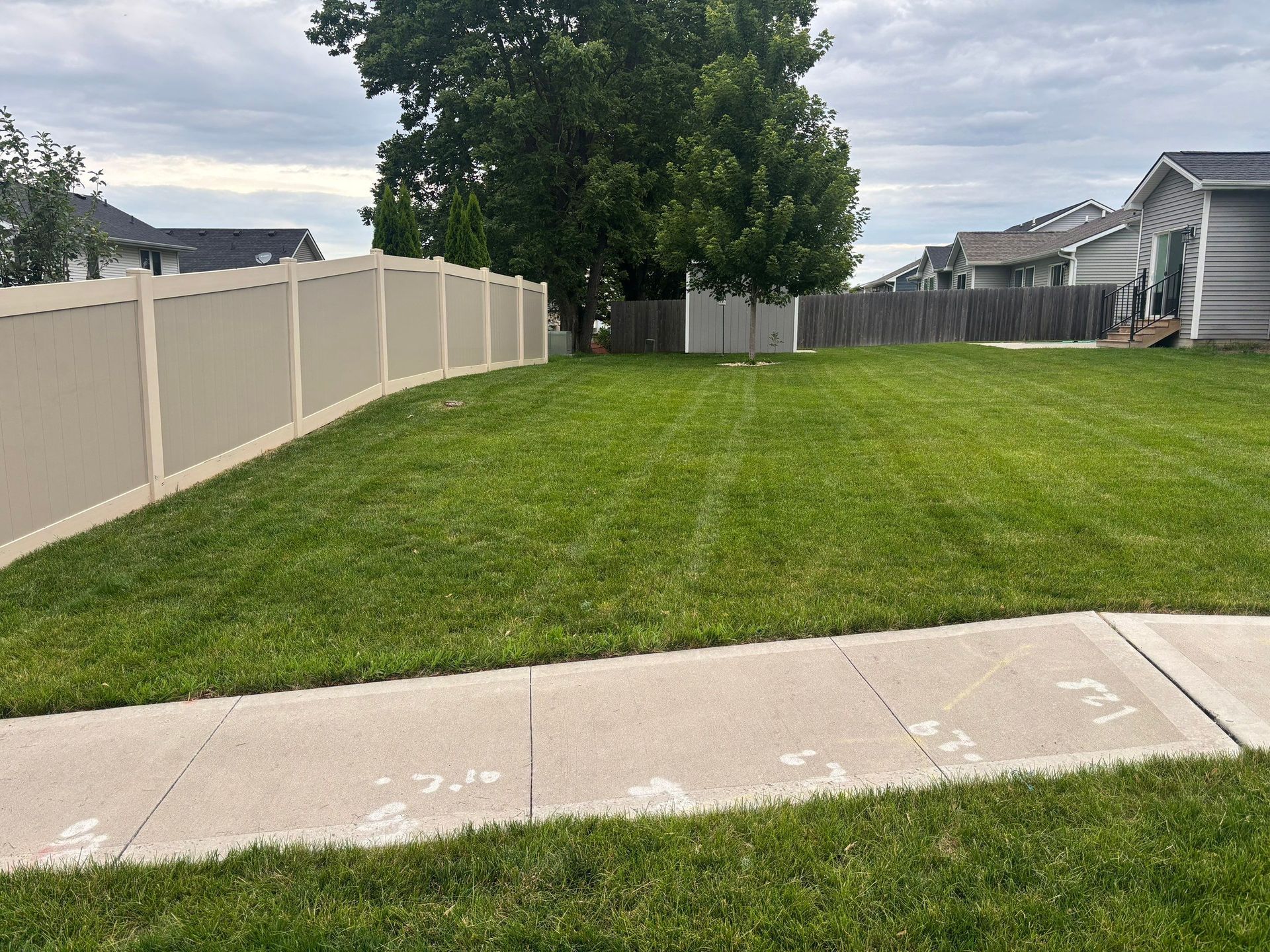 Suburban backyard with green lawn, white fence, and concrete sidewalk under a cloudy sky