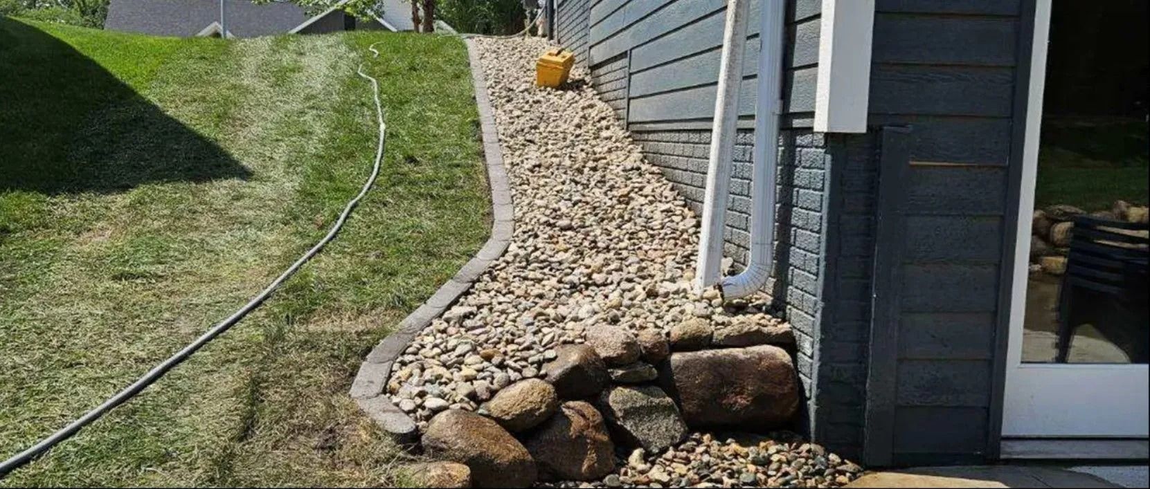 A gravel and rock border separates a grassy lawn from a gray house siding.