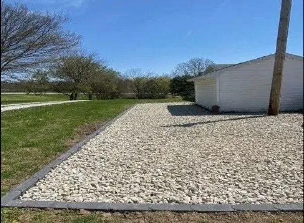 Gravel driveway bordered by concrete, next to a white building and grass field, under a blue sky.