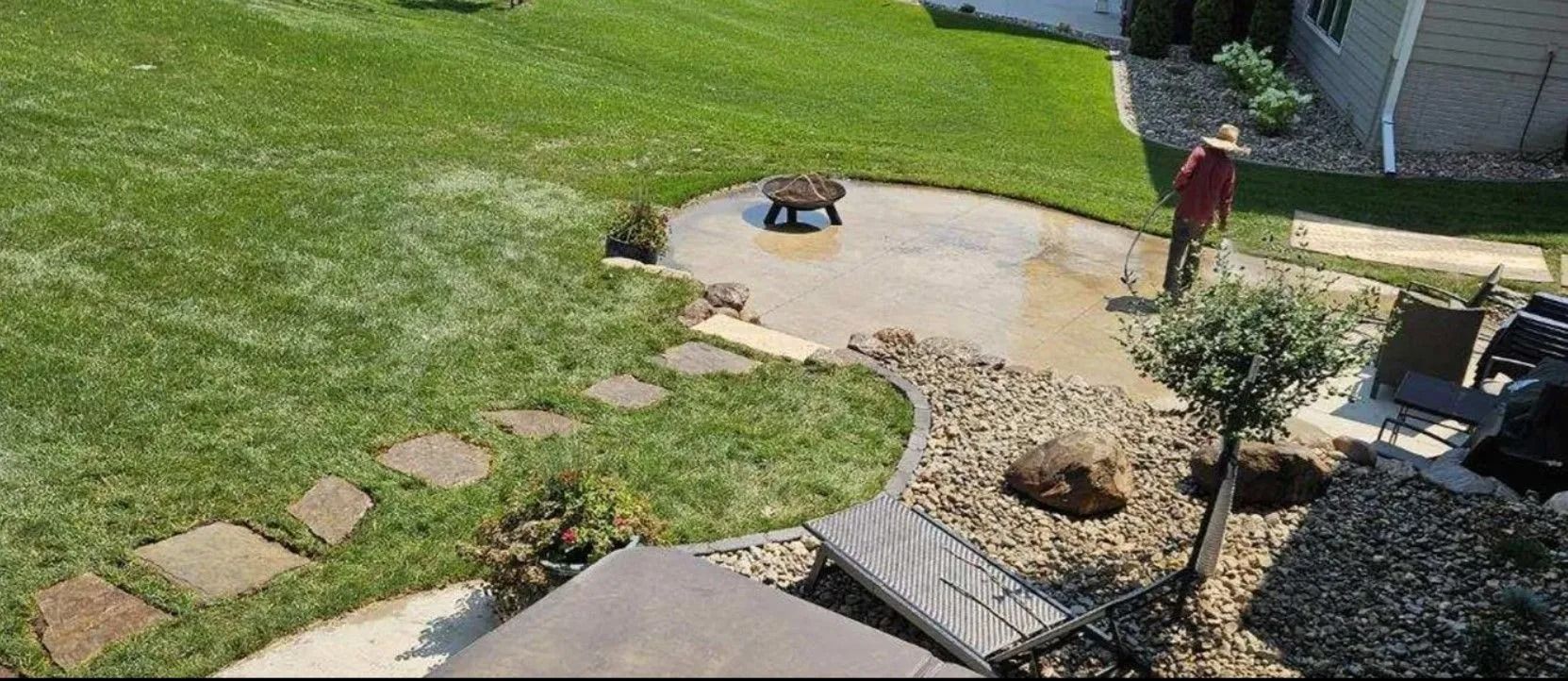 A person watering a backyard patio with a fire pit, stepping stones, and gravel path.