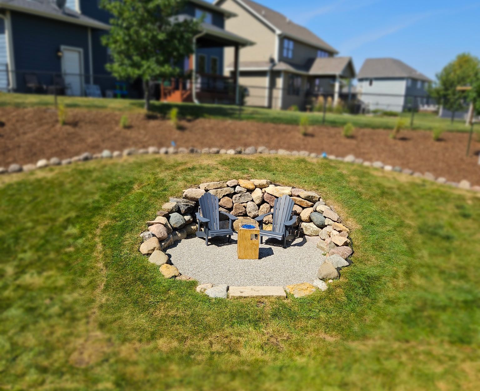 Fire pit area with two chairs, surrounded by rocks and gravel, in a grassy backyard.