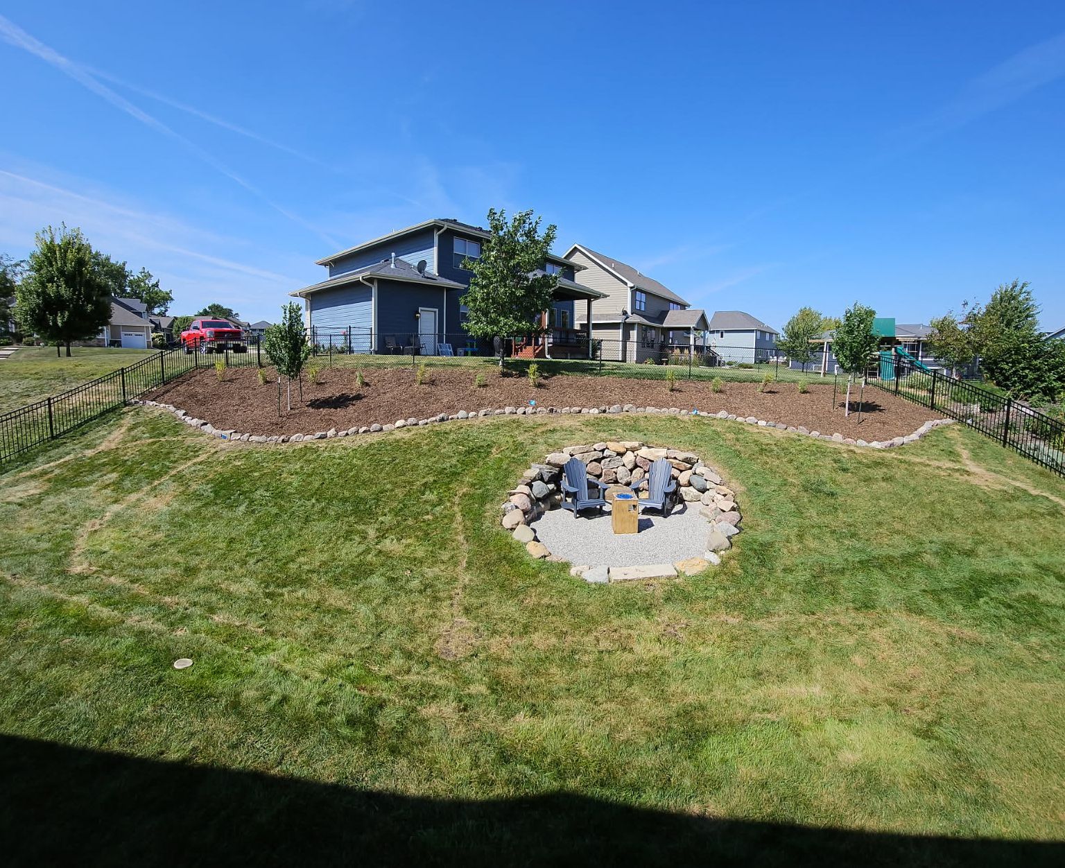Grassy backyard with a fire pit, tiered landscaping, and houses in the background under a blue sky.