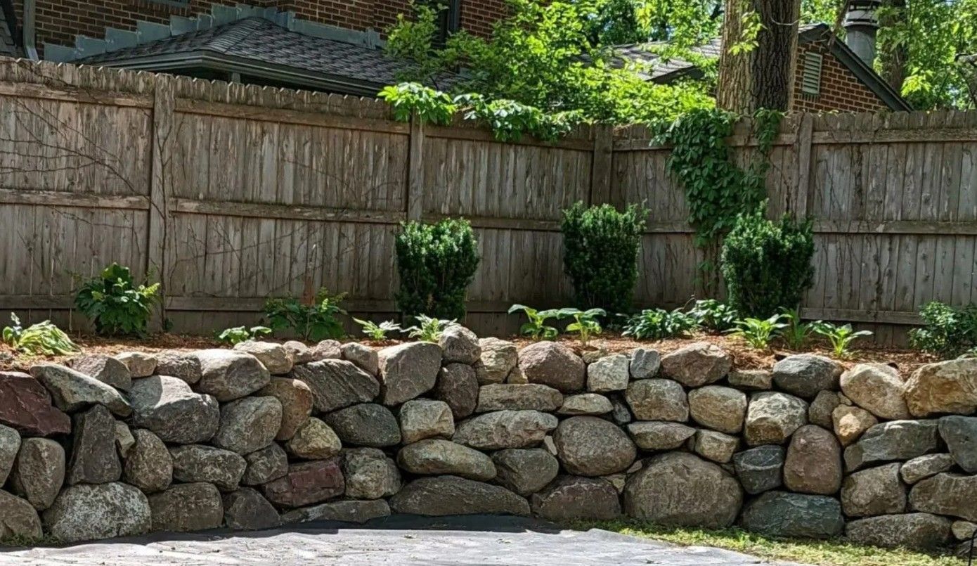 Stone retaining wall with a wooden fence and greenery behind it.