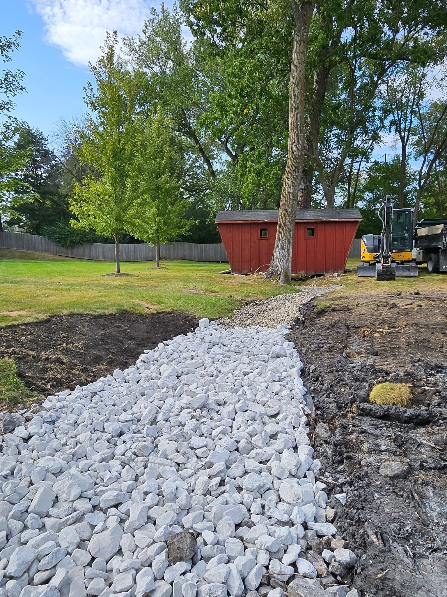 A rock-filled drainage channel in a grassy yard, with a red shed and trees in the background.