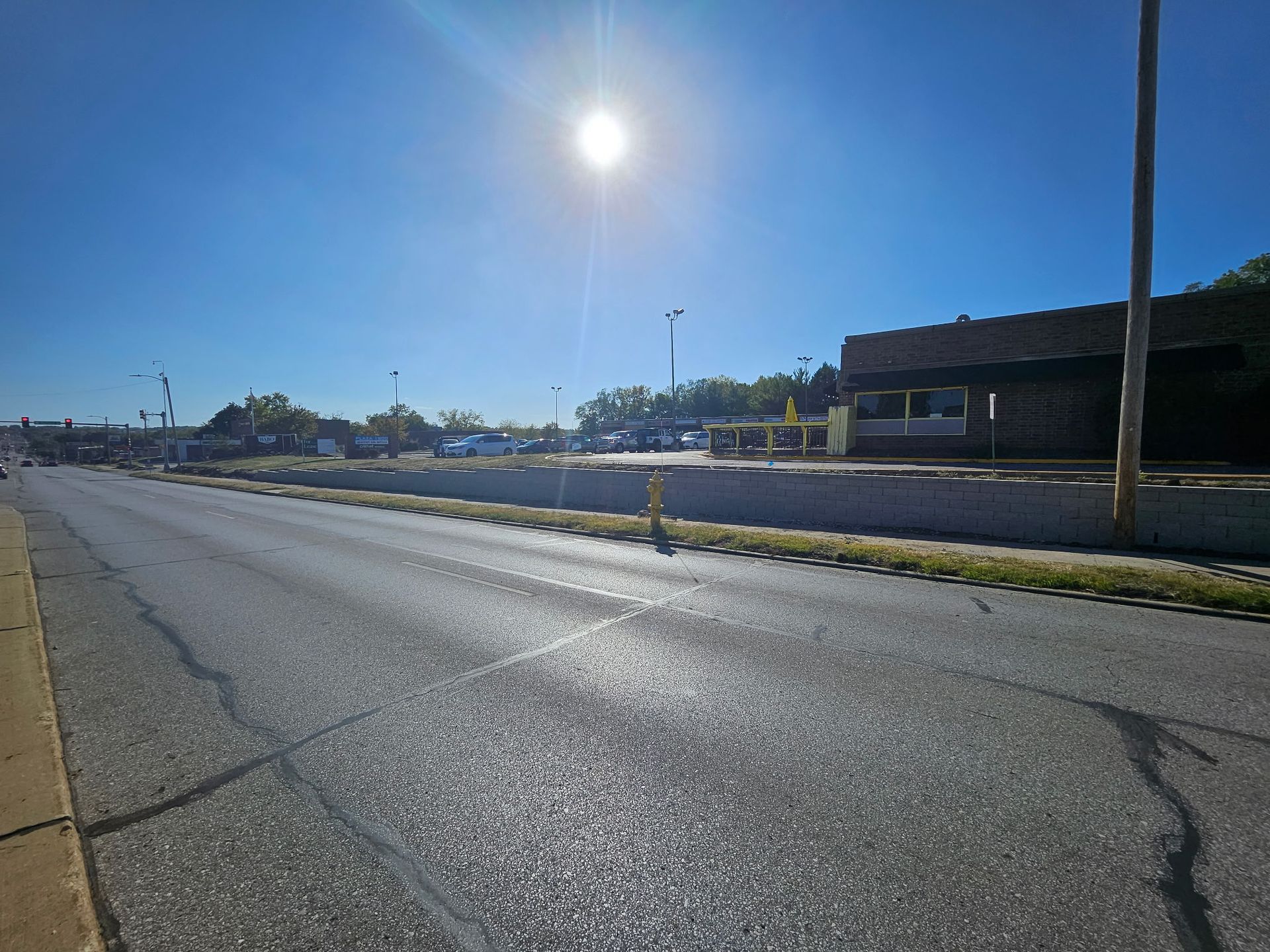 Sunlit asphalt road, low concrete barrier with weeds, building with dark facade, bright blue sky.