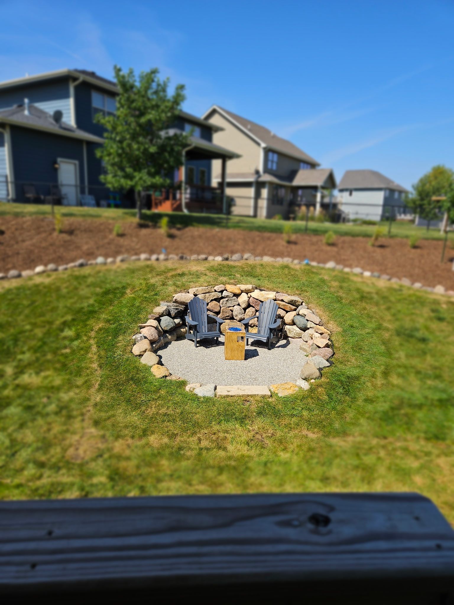 Fire pit with two chairs in a backyard, surrounded by stone and grass. Houses and blue sky in background.