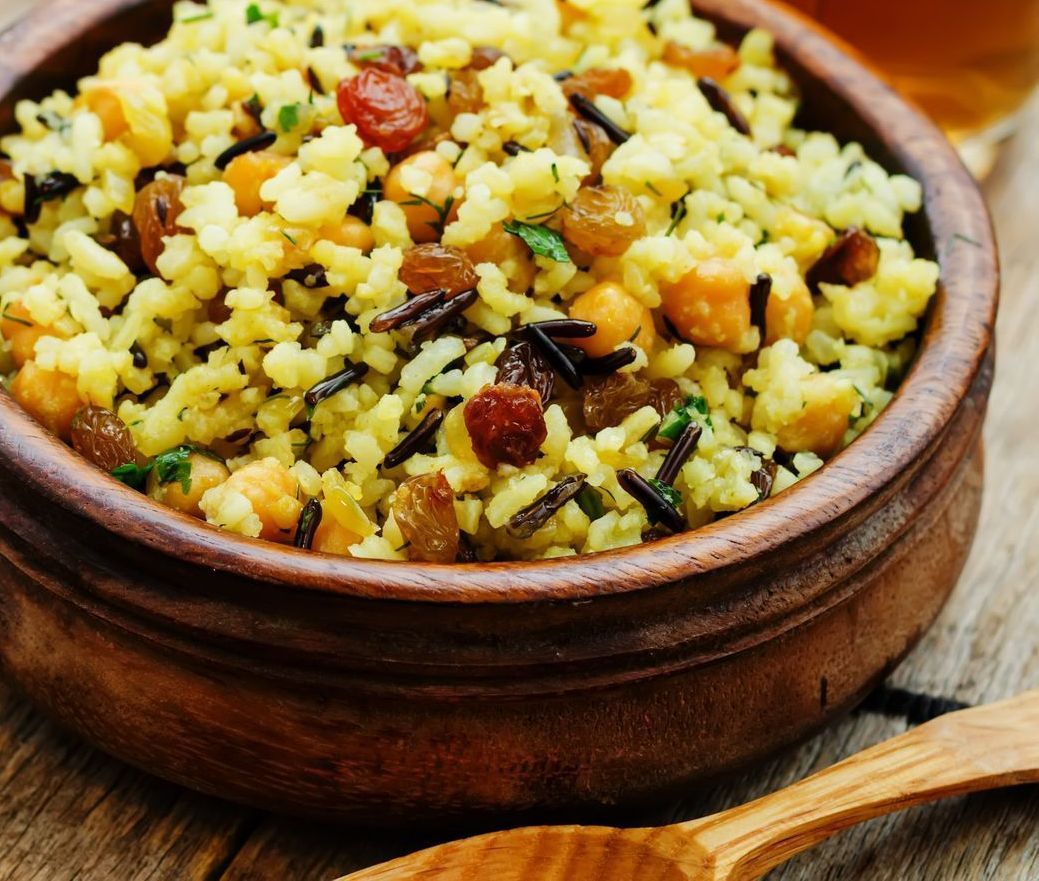 Wooden bowl of yellow rice pilaf with herbs, nuts, and raisins on a rustic table
