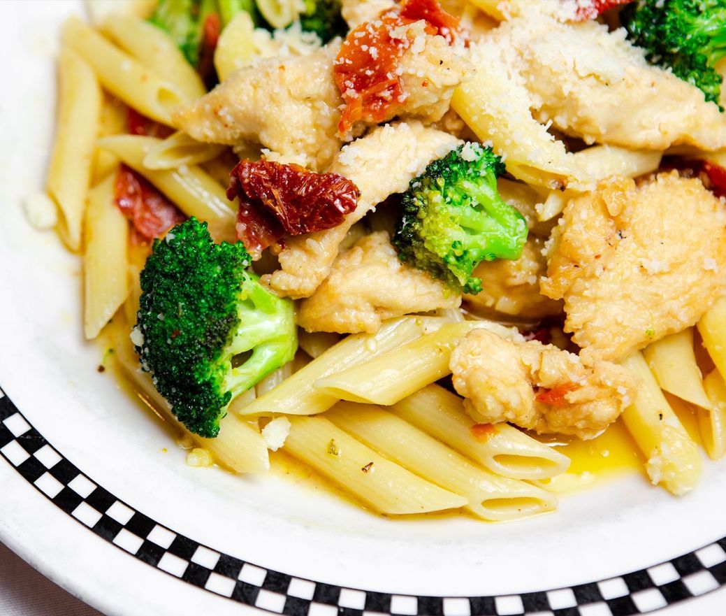 Pasta with chicken, broccoli, and sun-dried tomatoes in a white bowl with a black-checkered rim