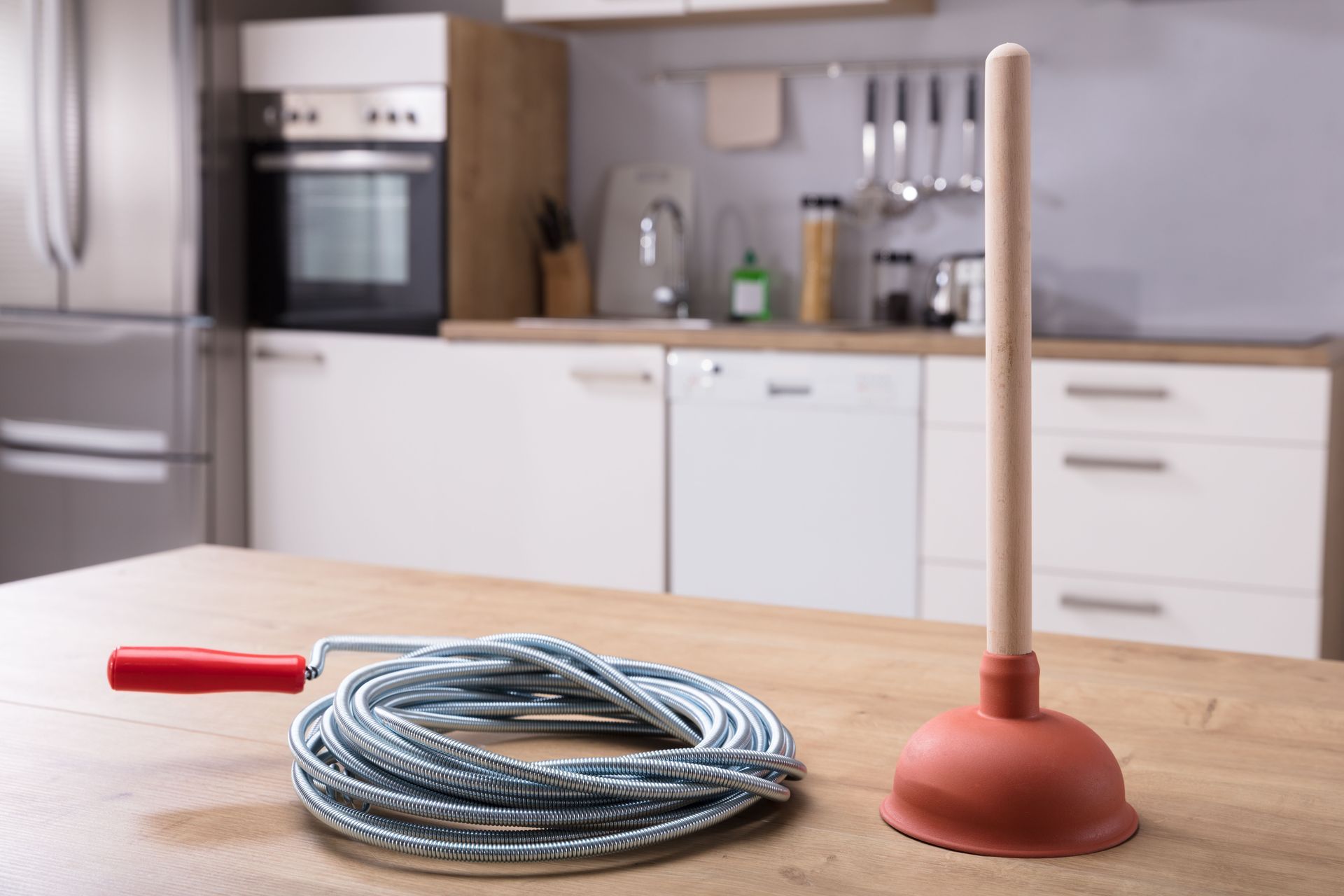 Plunger and plumbing snake on a kitchen counter in front of cabinetry and appliances.
