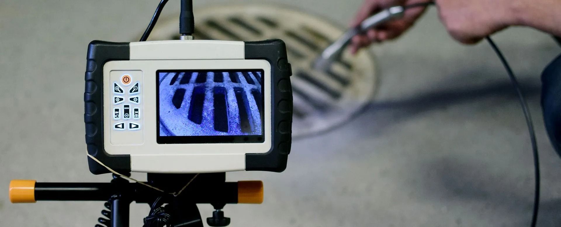 Camera inspecting a sewer grate, displaying the inside. Person holding a tool, on a light grey concrete surface.