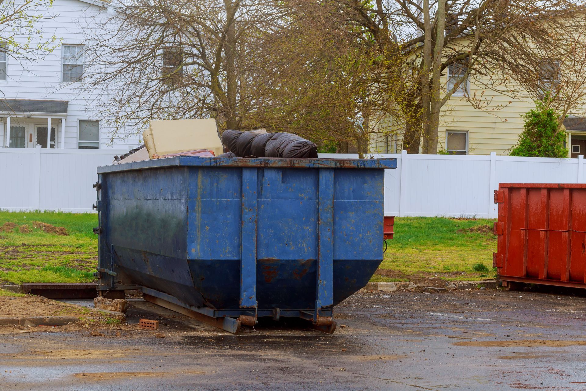 A blue dumpster partially filled with debris sits on an asphalt lot in front of houses and a white fence.