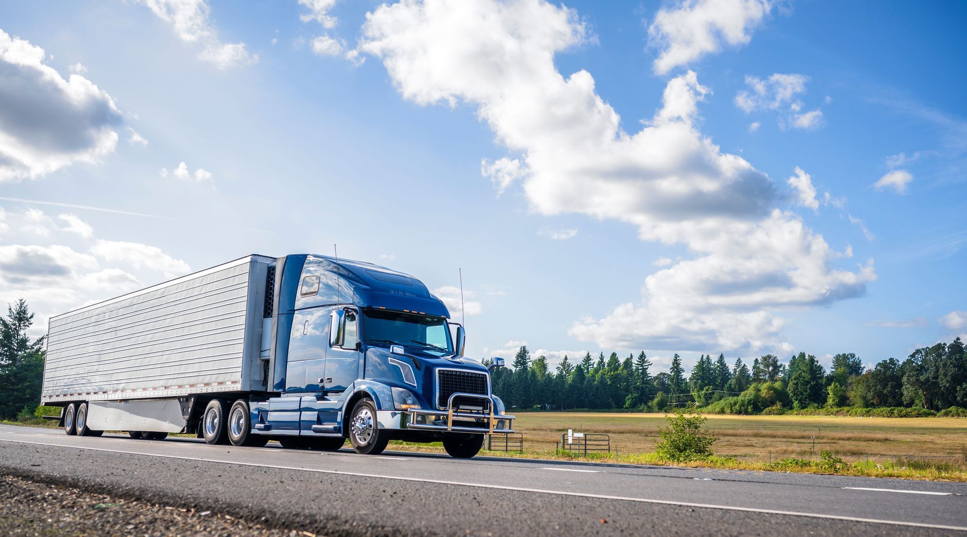 Blue semi-truck driving on a road under a partly cloudy blue sky.