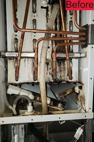 Close-up of a dirty, open boiler with copper pipes, a white plastic tube, and black residue.