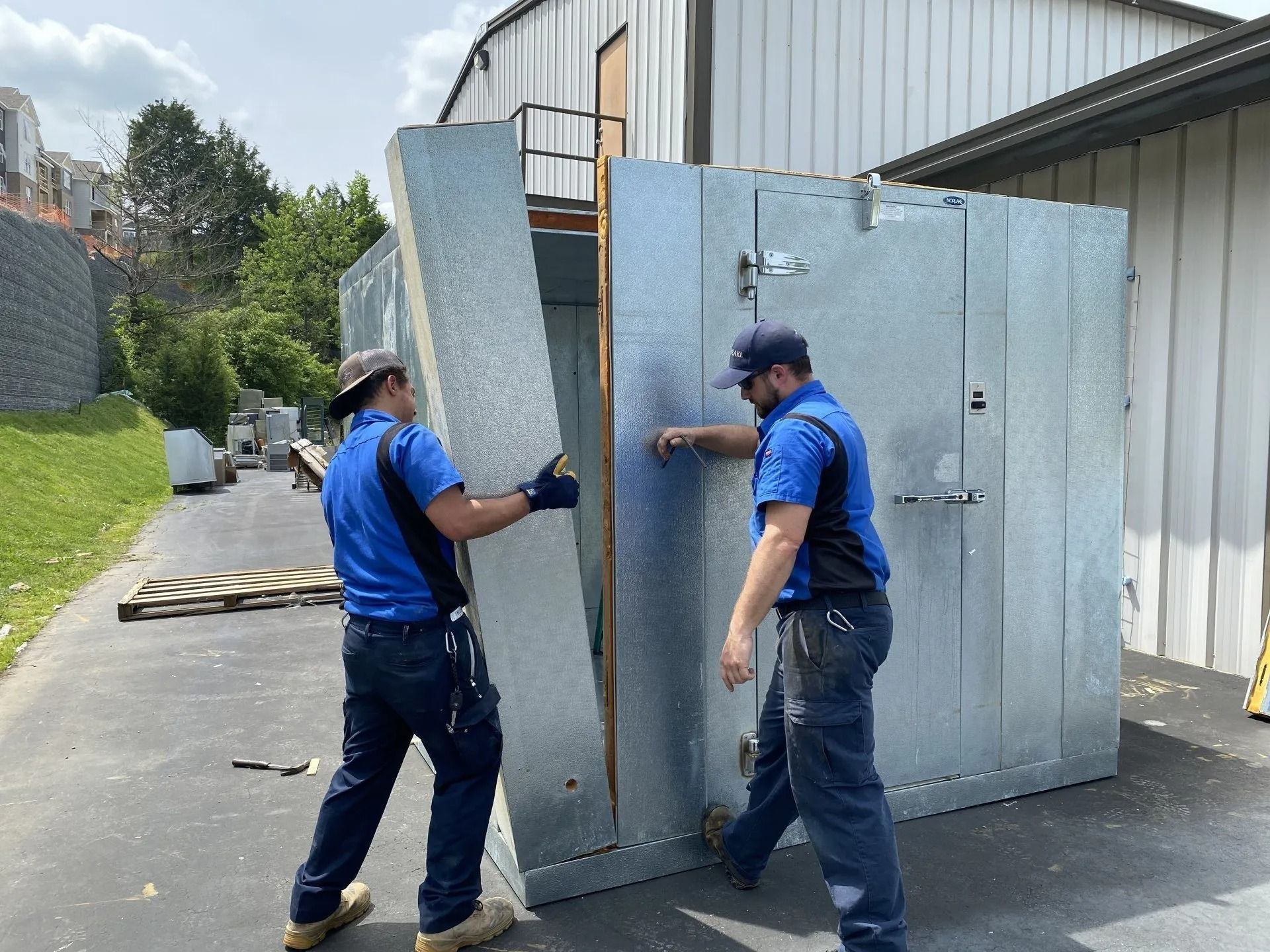 Two workers installing a large, metallic walk-in cooler outside a building.