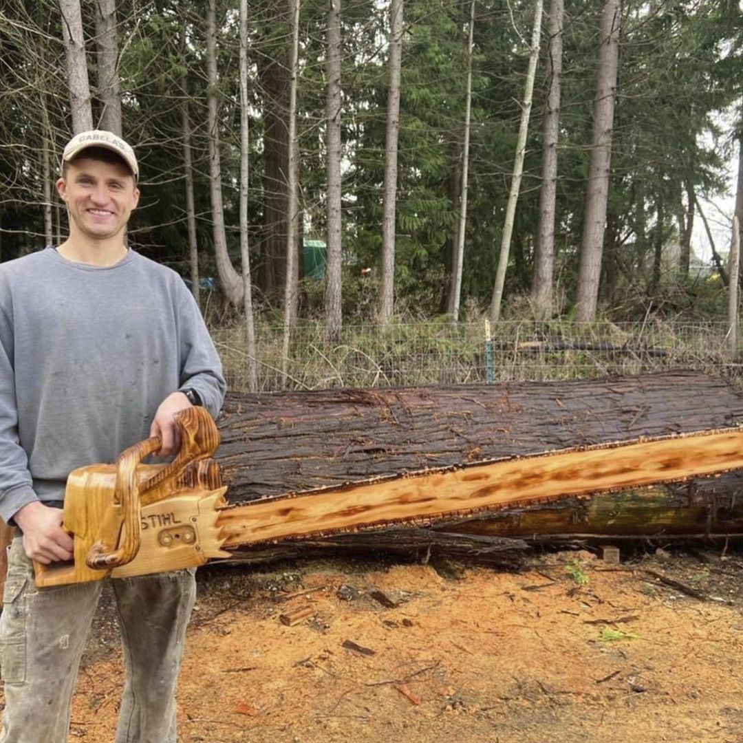Man holding wooden chainsaw carving, standing in front of a carved log in a wooded area.