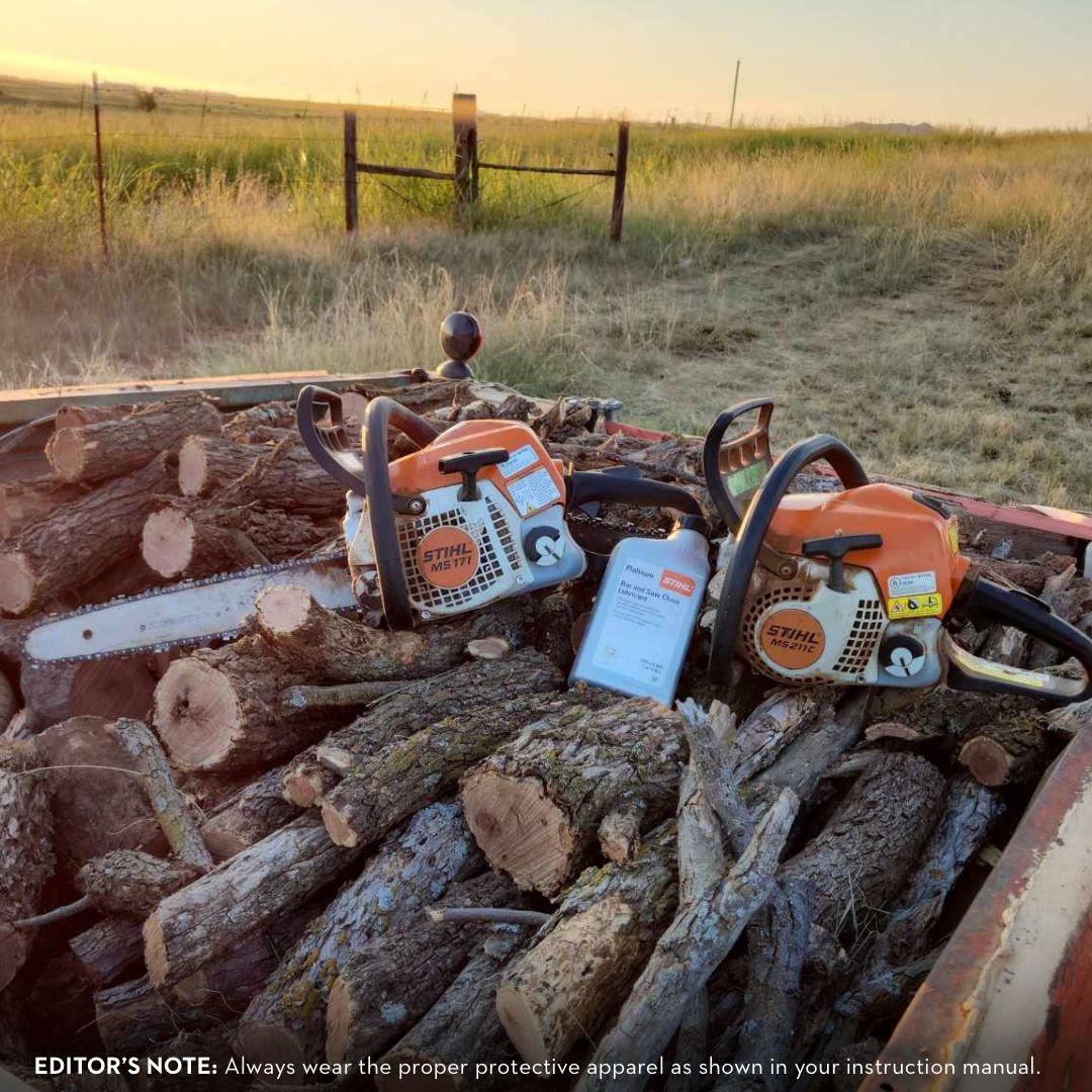 Two orange Stihl chainsaws on a pile of logs, with a bottle of oil in a field at sunset.