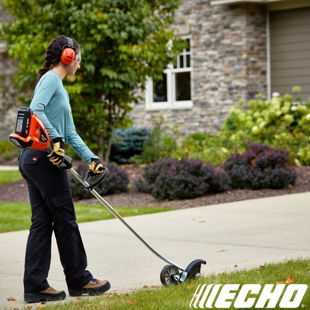 Woman using a string trimmer to edge a sidewalk next to a house with shrubs.