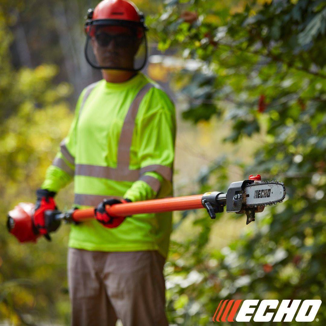 Person using a pole saw, wearing safety gear, outdoors. The saw is orange and black.