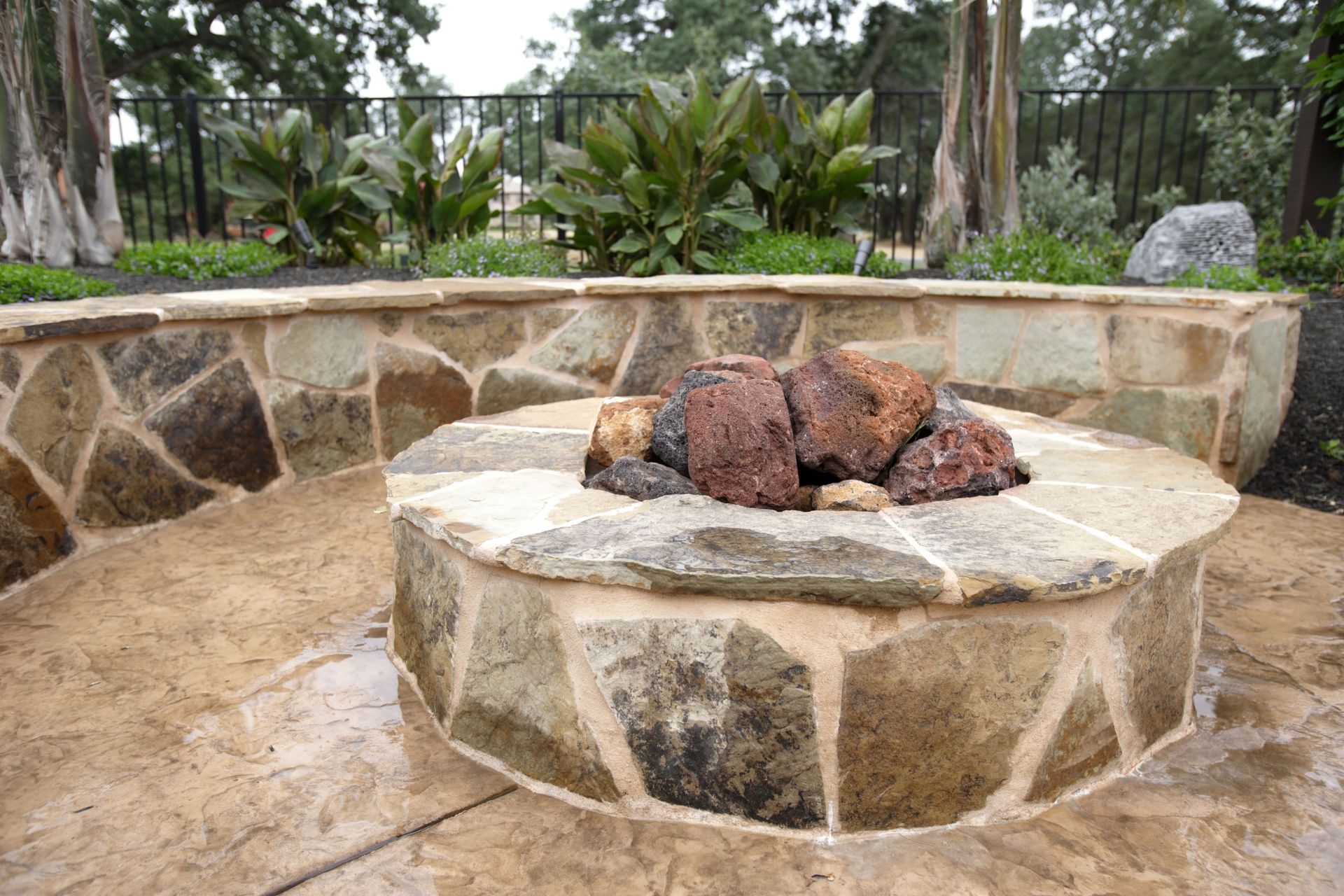 Stone fire pit with seating in a garden setting, dark rocks in pit, surrounding stone patio.