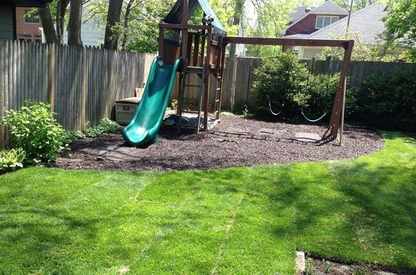 Playground set with green slide and swings in a backyard with a wooden fence and lawn.
