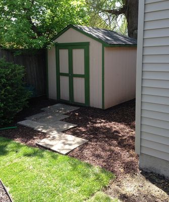 Small tan shed with green trim in a backyard setting, surrounded by mulch and stepping stones.
