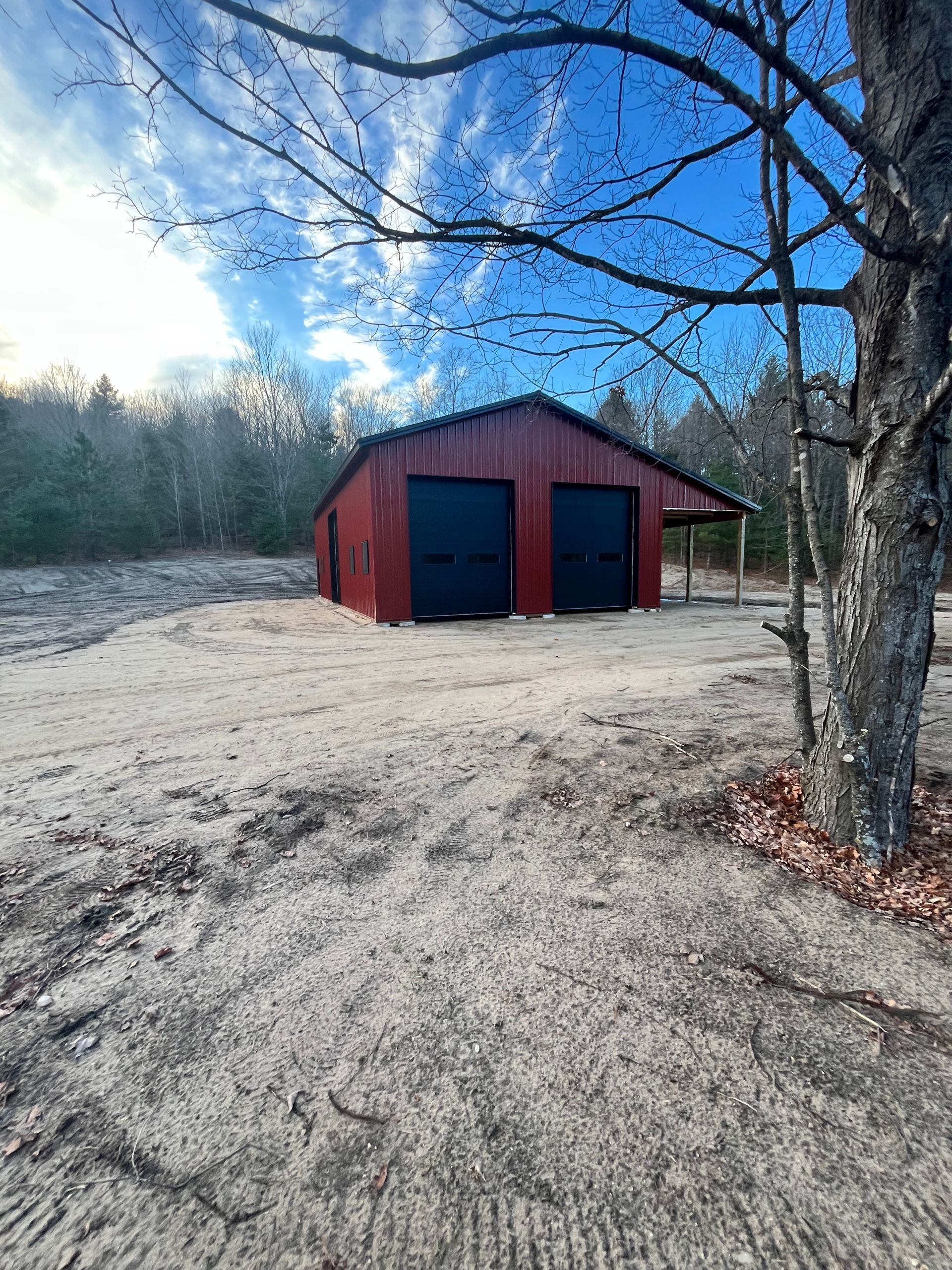 Red barn with black garage doors on a sandy lot, under a blue sky with tree branches.