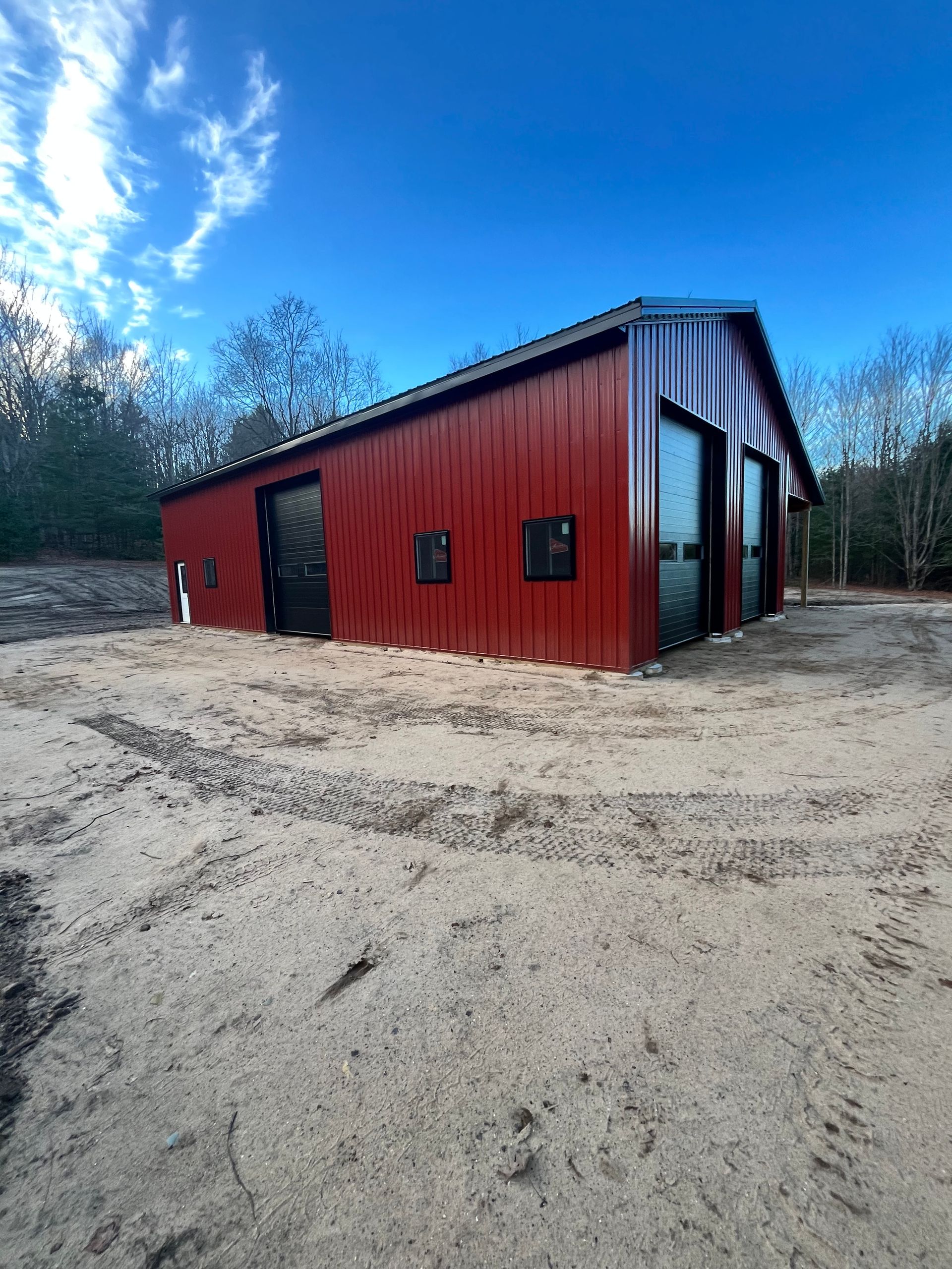 Red metal barn with black doors and windows against a blue sky, set on a sandy lot.