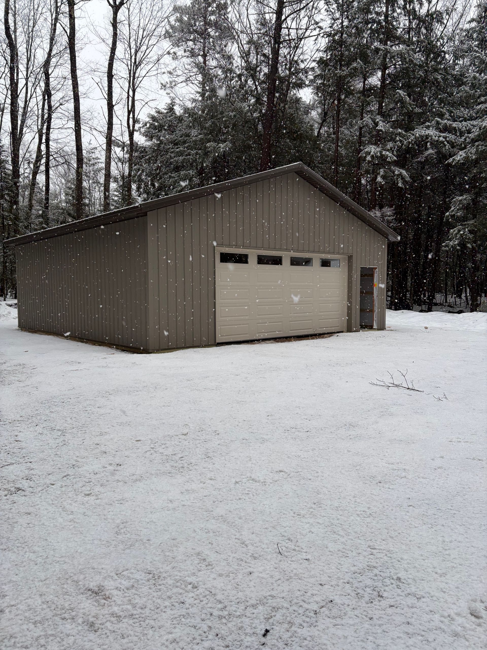 Snow-covered ground and a metal-sided garage in a wooded area during a snowfall.