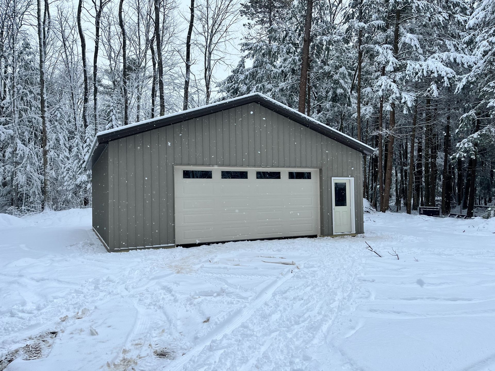 A grey metal garage with a white door and side entry sits in a snowy, wooded setting.
