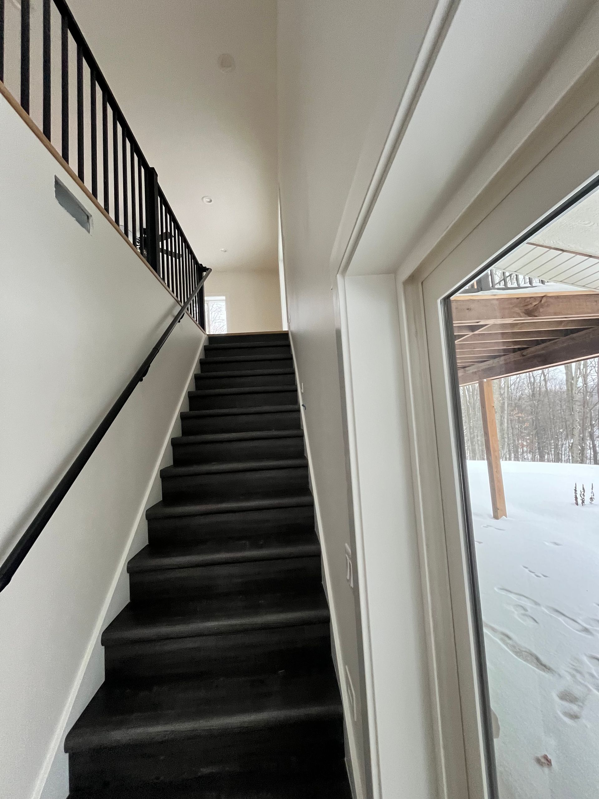 Staircase with black carpet, black railing, leading up to a hallway. Snow seen outside window.