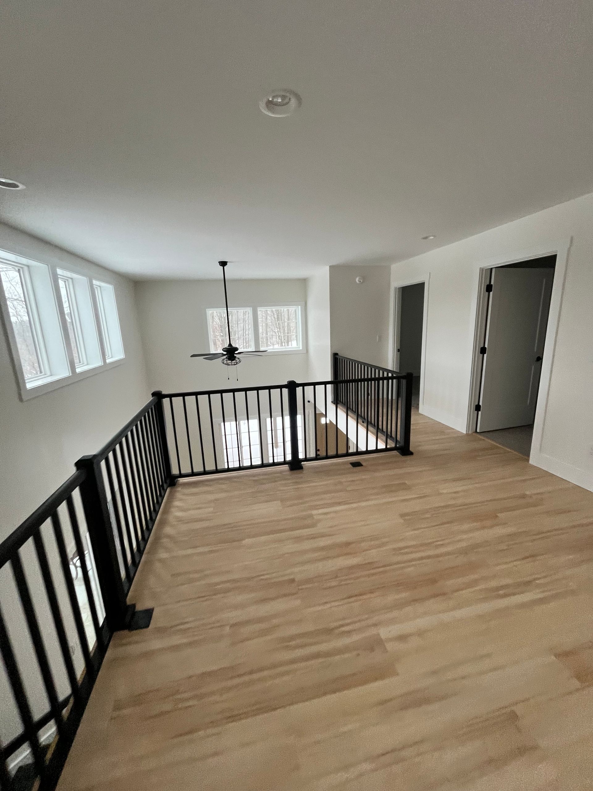 Interior view of a loft with light wood floors, black railing, and white walls. Windows and a ceiling fan are visible.