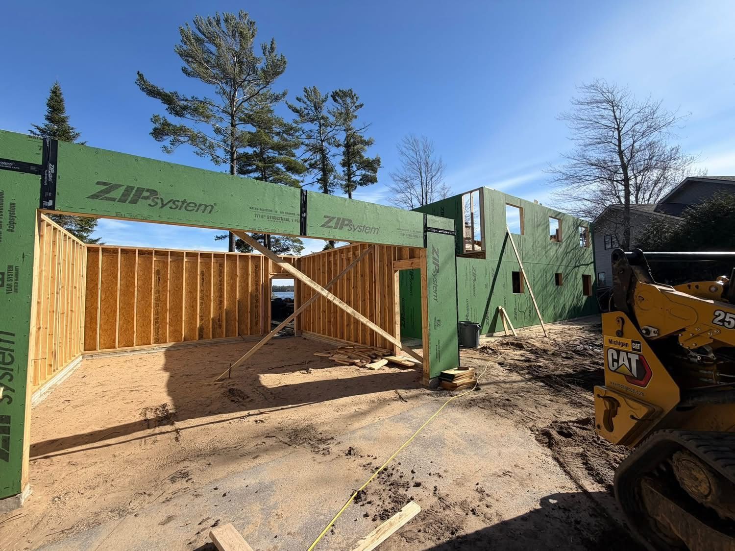 Green house framing under construction with plywood walls, blue sky, and a skid steer at right