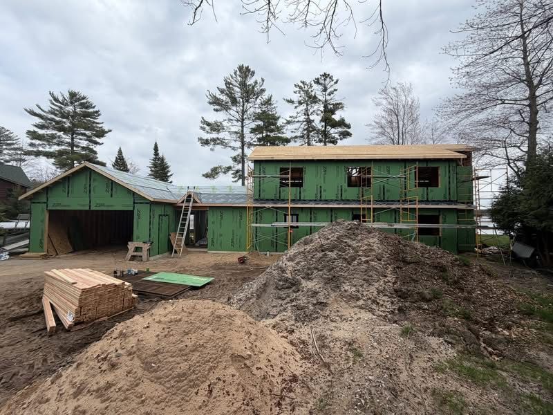 Green two-story house under construction with piles of dirt and lumber in a wooded area
