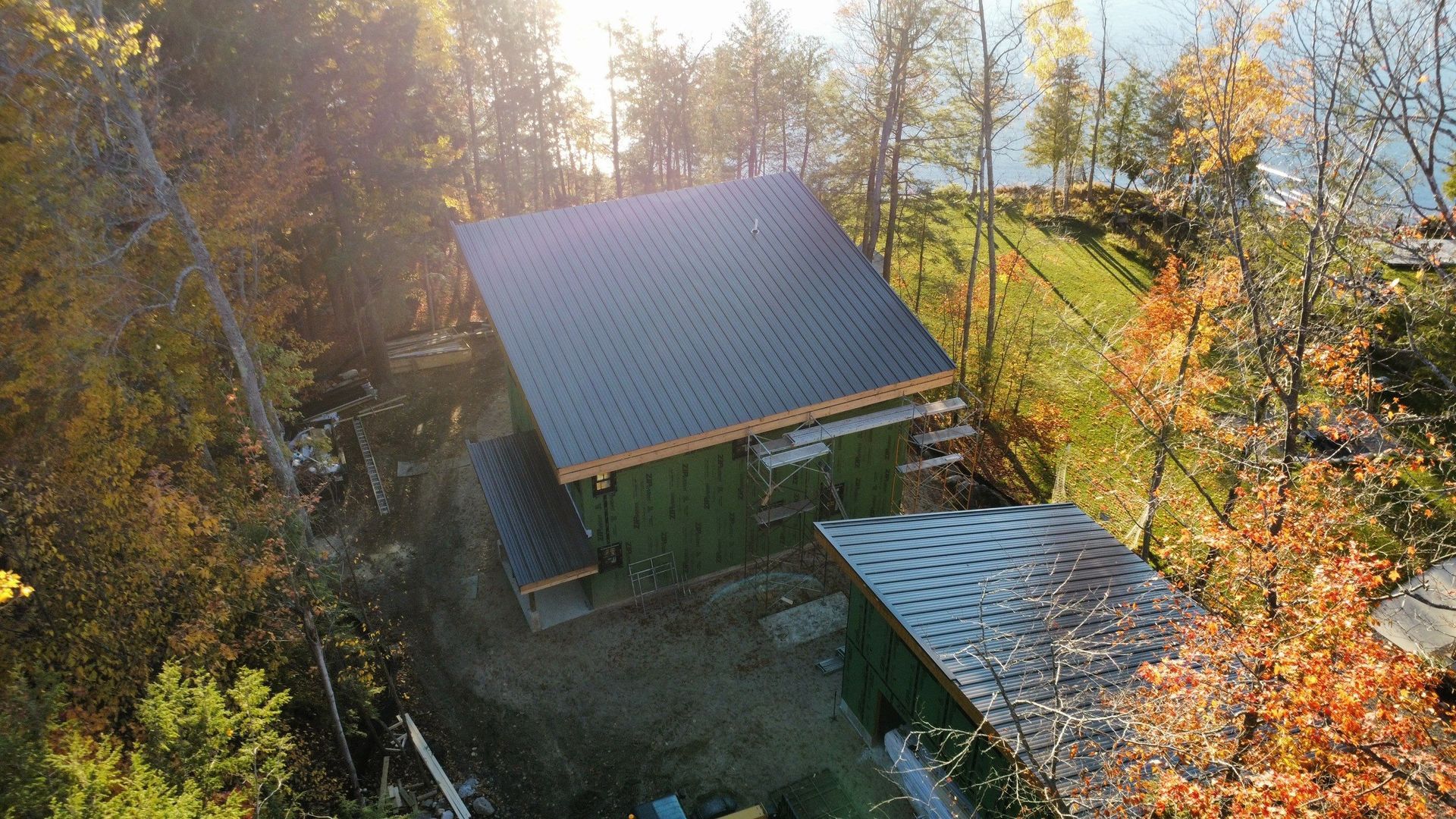 Green-sided house with dark roof surrounded by trees and overlooking a body of water.