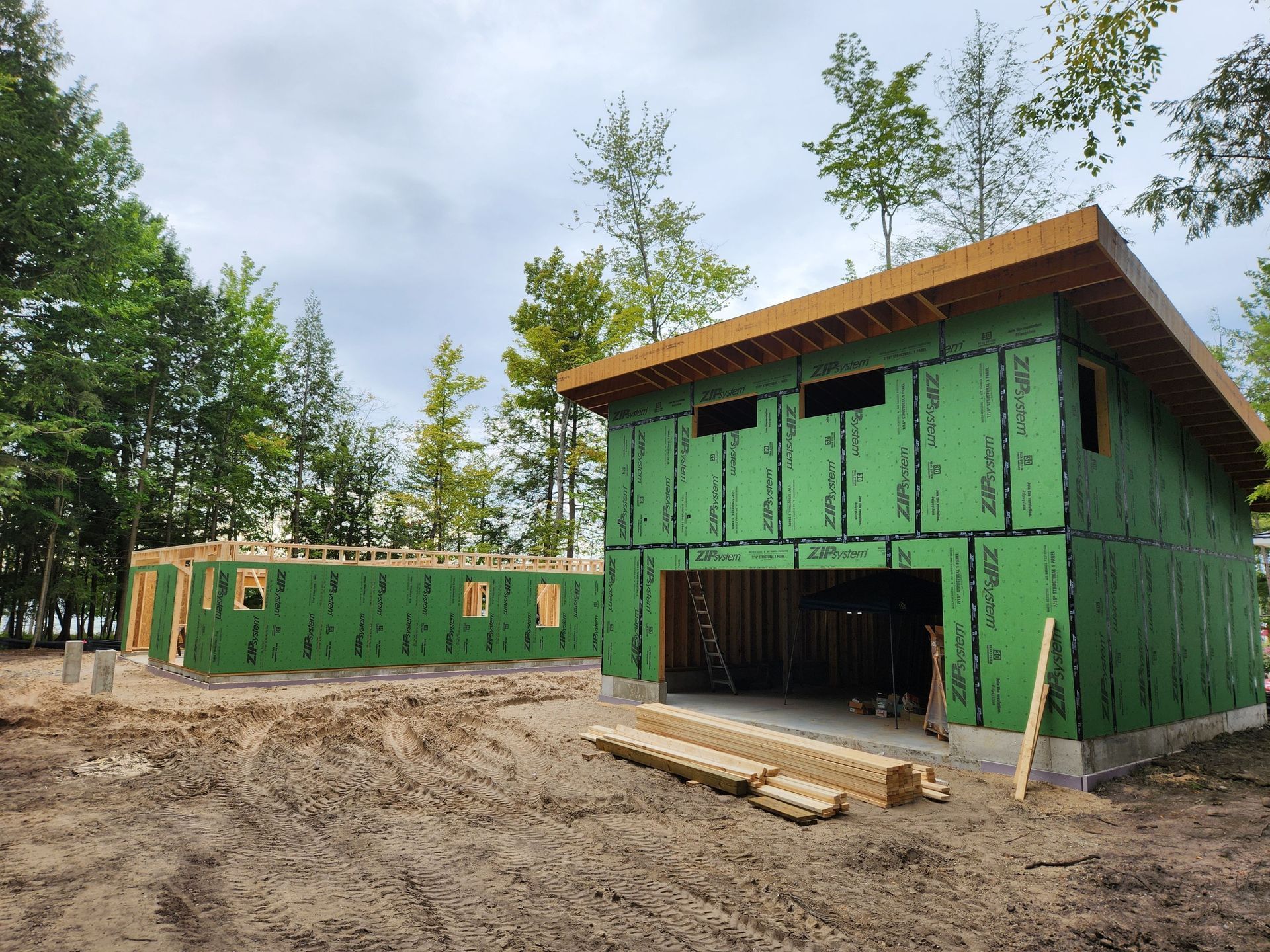 Two-story building under construction with green sheathing and open garage door, trees in the background.