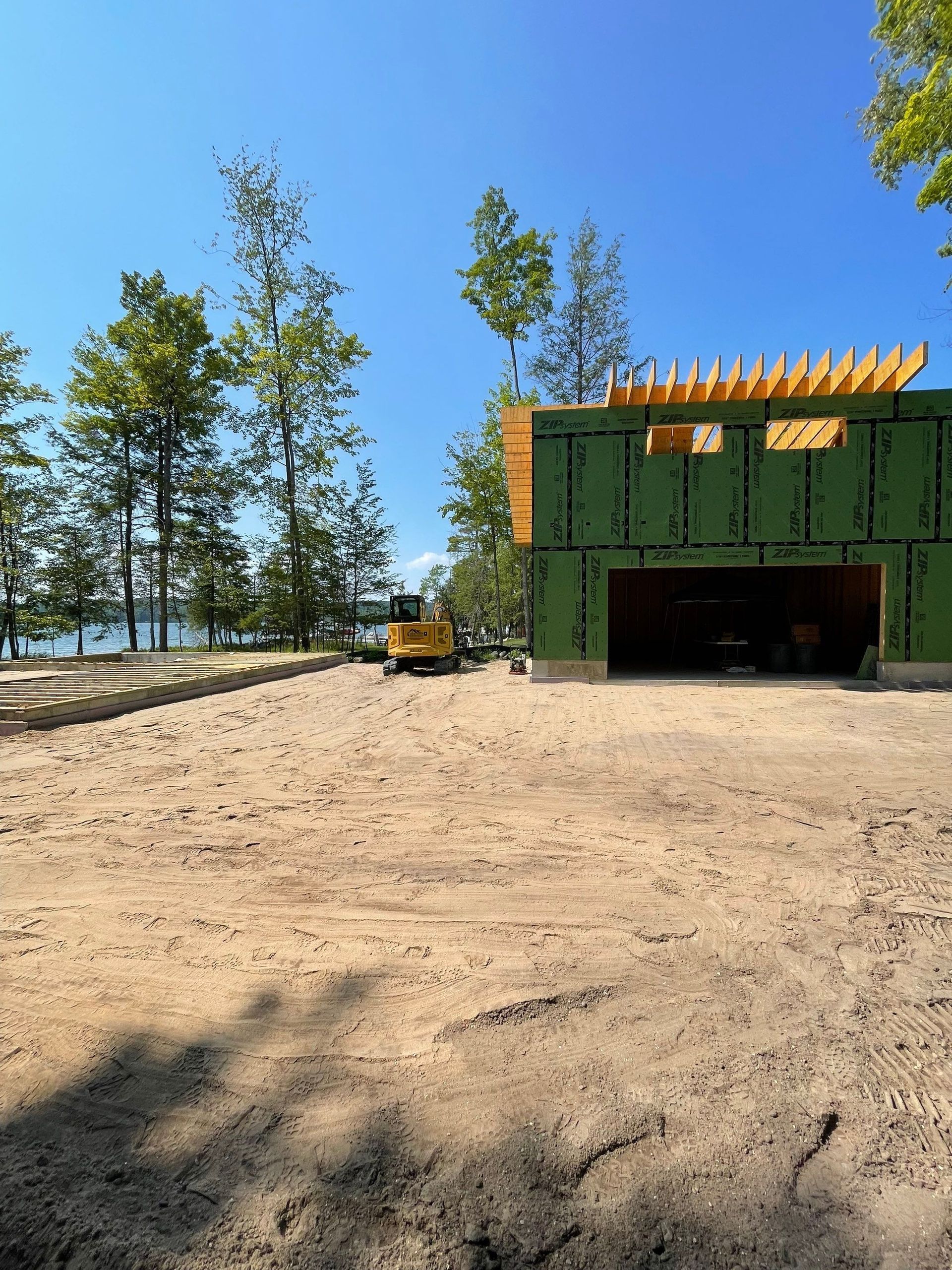 Construction site with a partially built green-sided building, excavator, and dirt lot, trees and water in the background.
