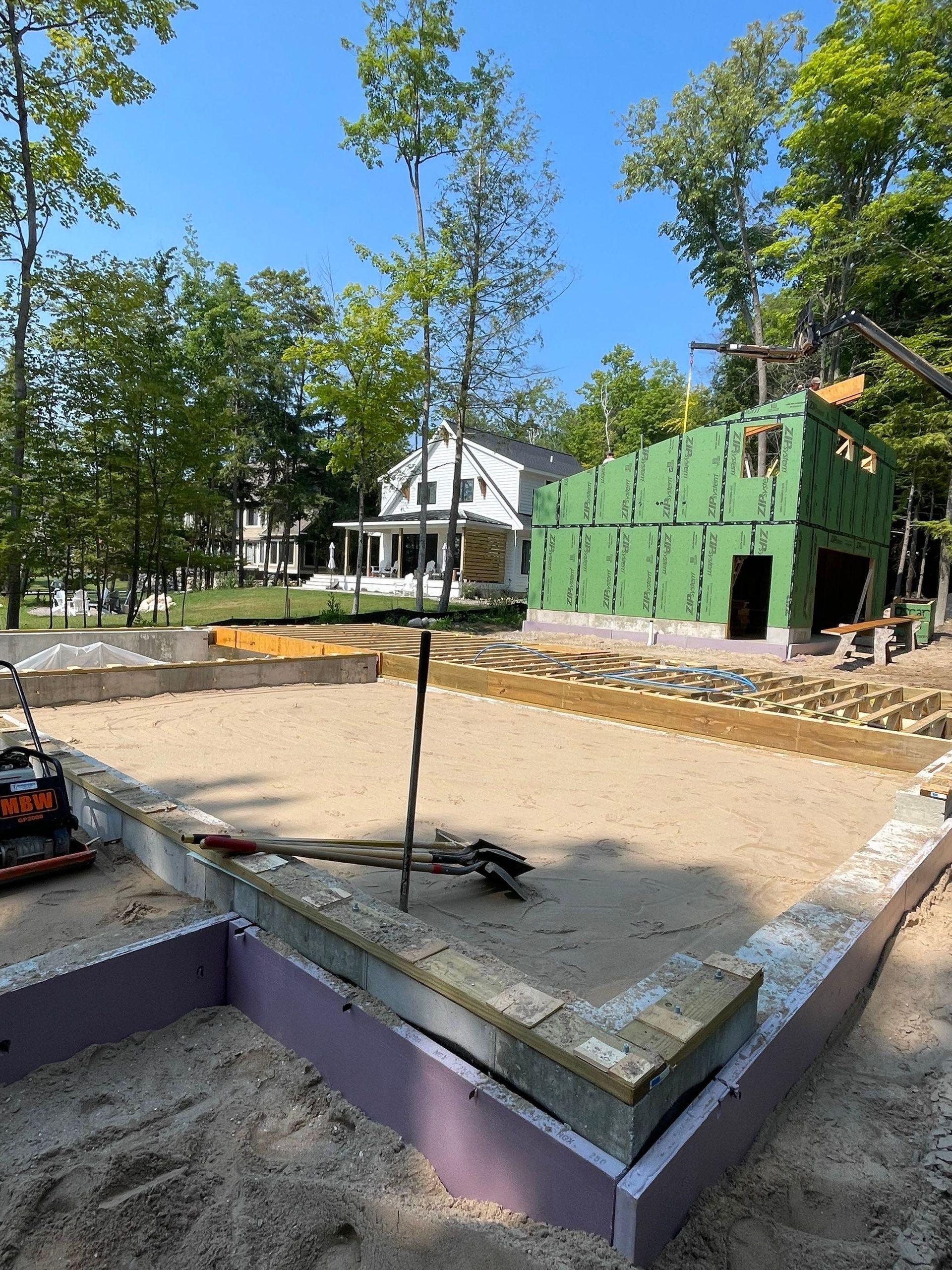 Construction site: a building's foundation and partially built structure. Green sheathing, sand, and trees in background.