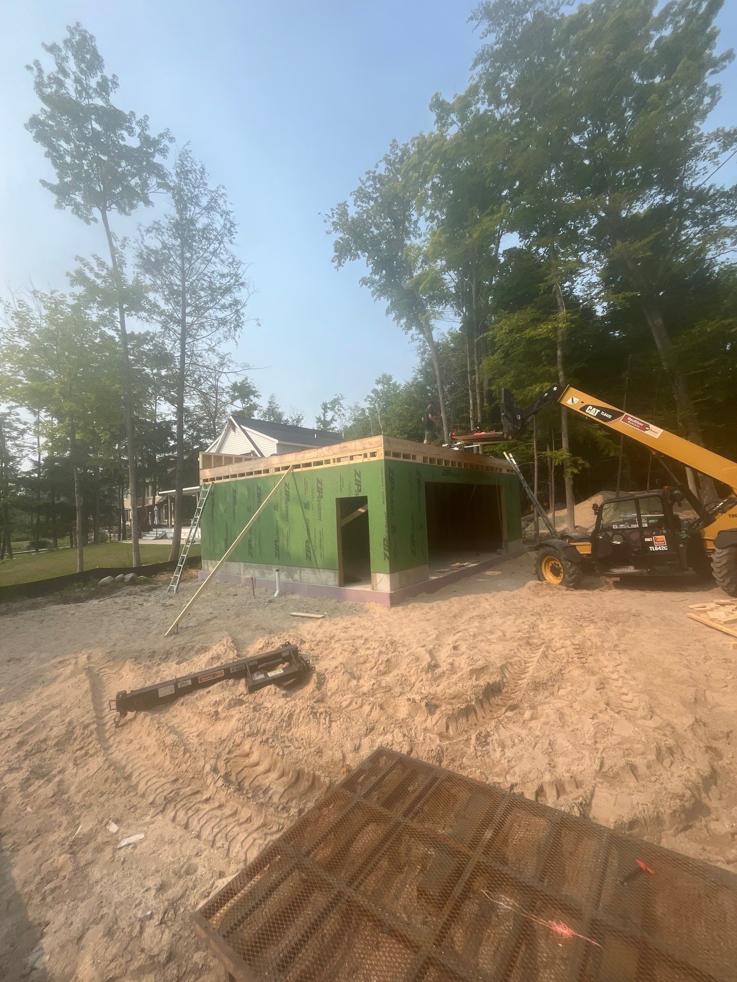 Garage under construction with green plywood walls and wood framing, trees in the background, bright sunlight.