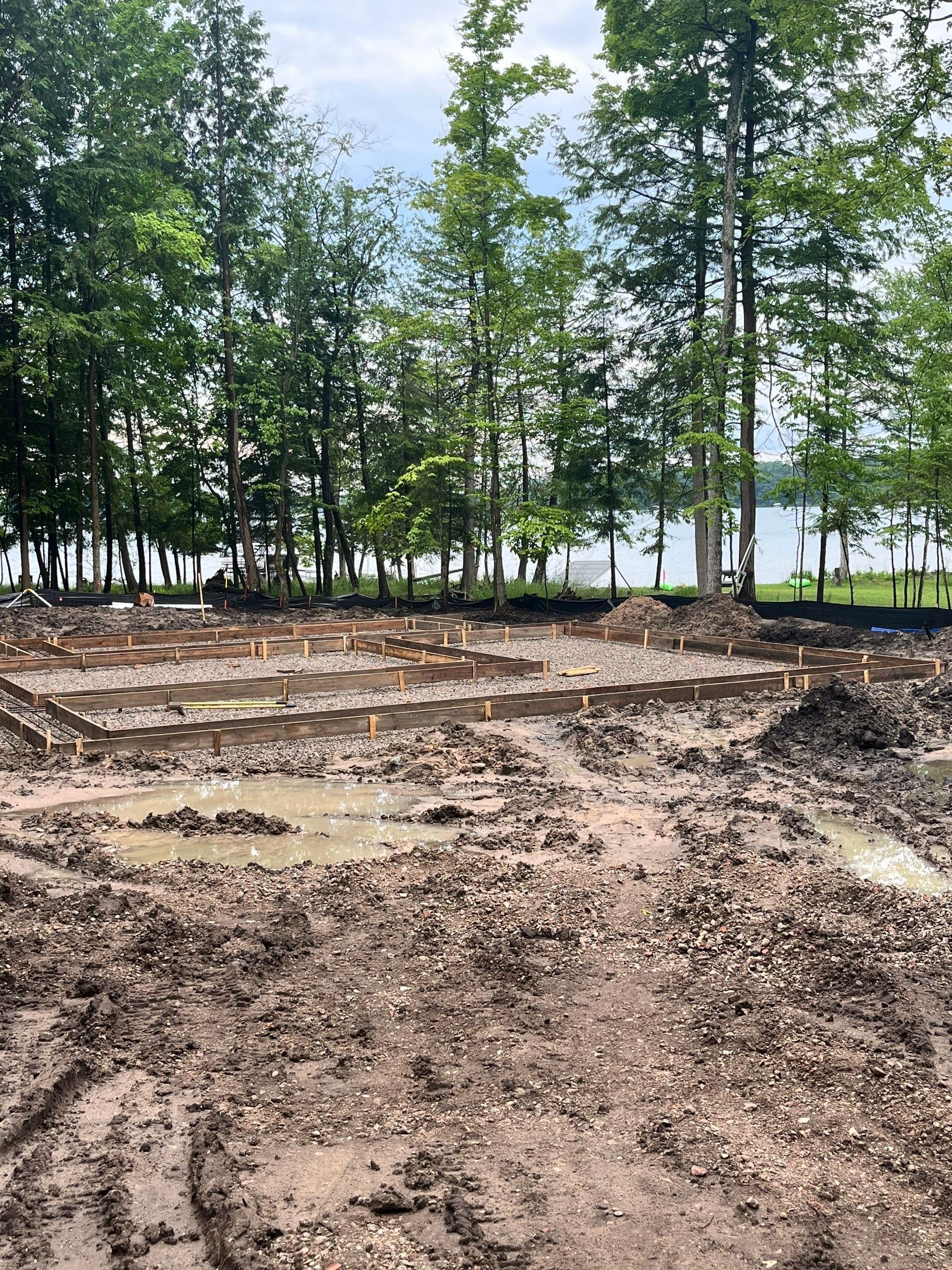 Construction site: excavated muddy ground with wooden framework, trees and water in background.