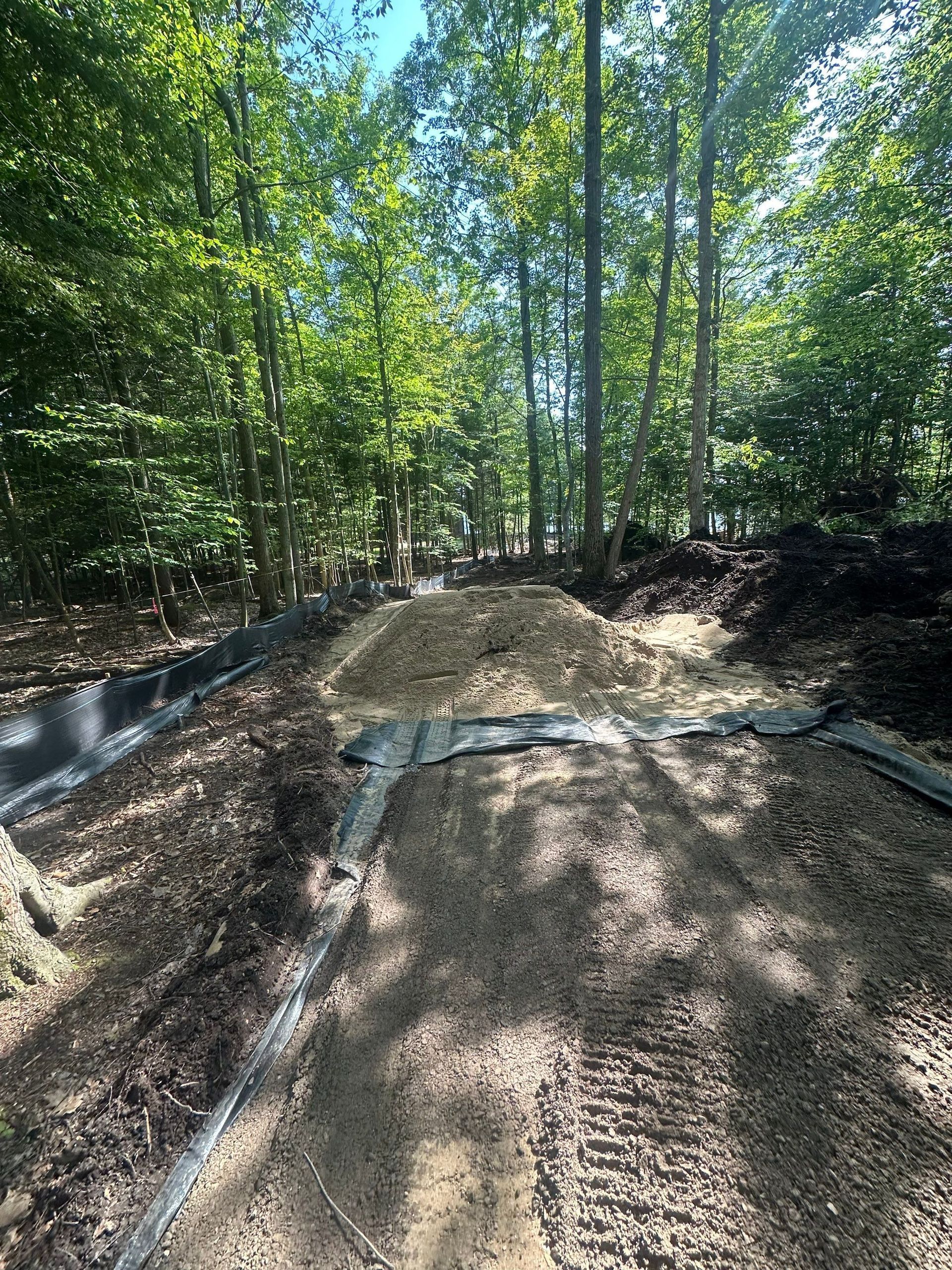 Dirt path with raised edges, surrounded by trees and mulch piles.