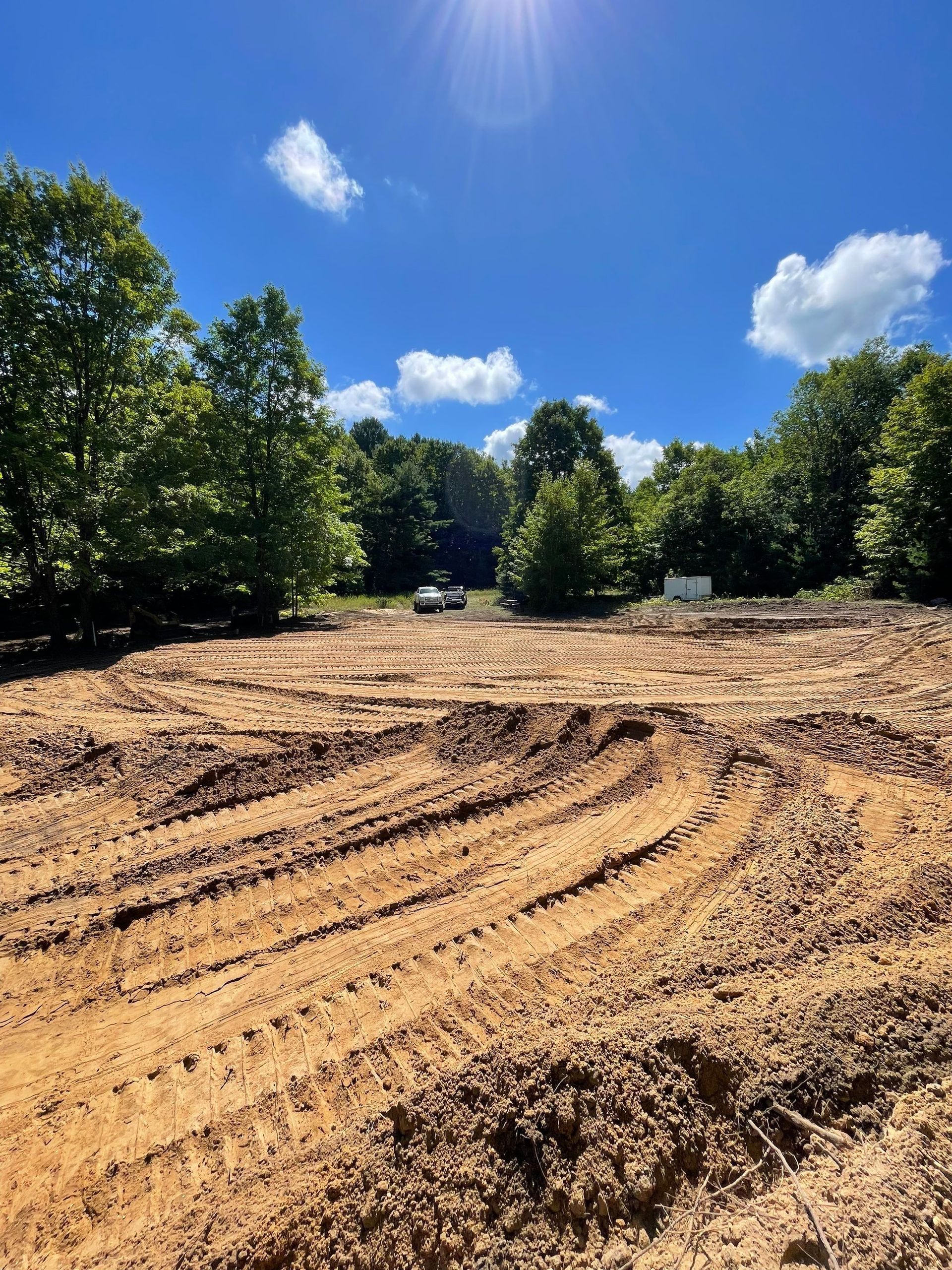 Cleared land with tire tracks under a sunny blue sky, surrounded by trees.