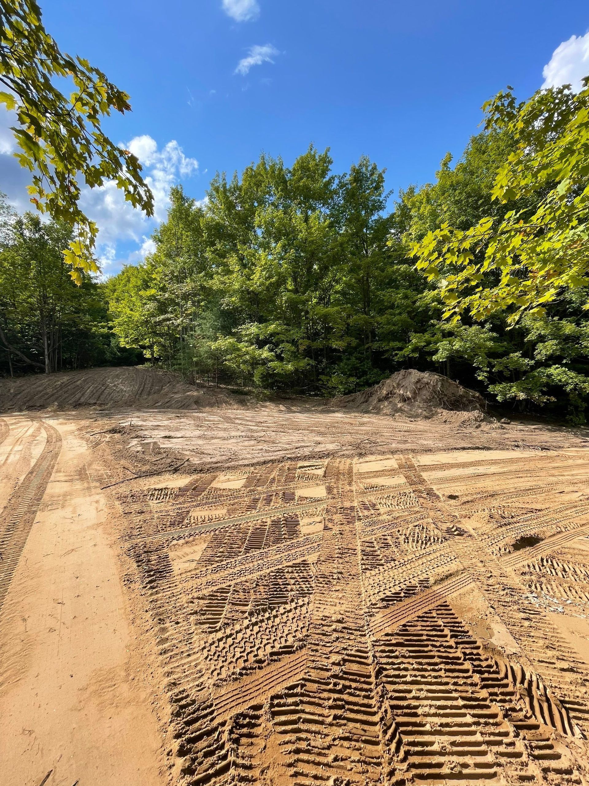 Dirt path with tire tracks in front of green trees under a blue sky.