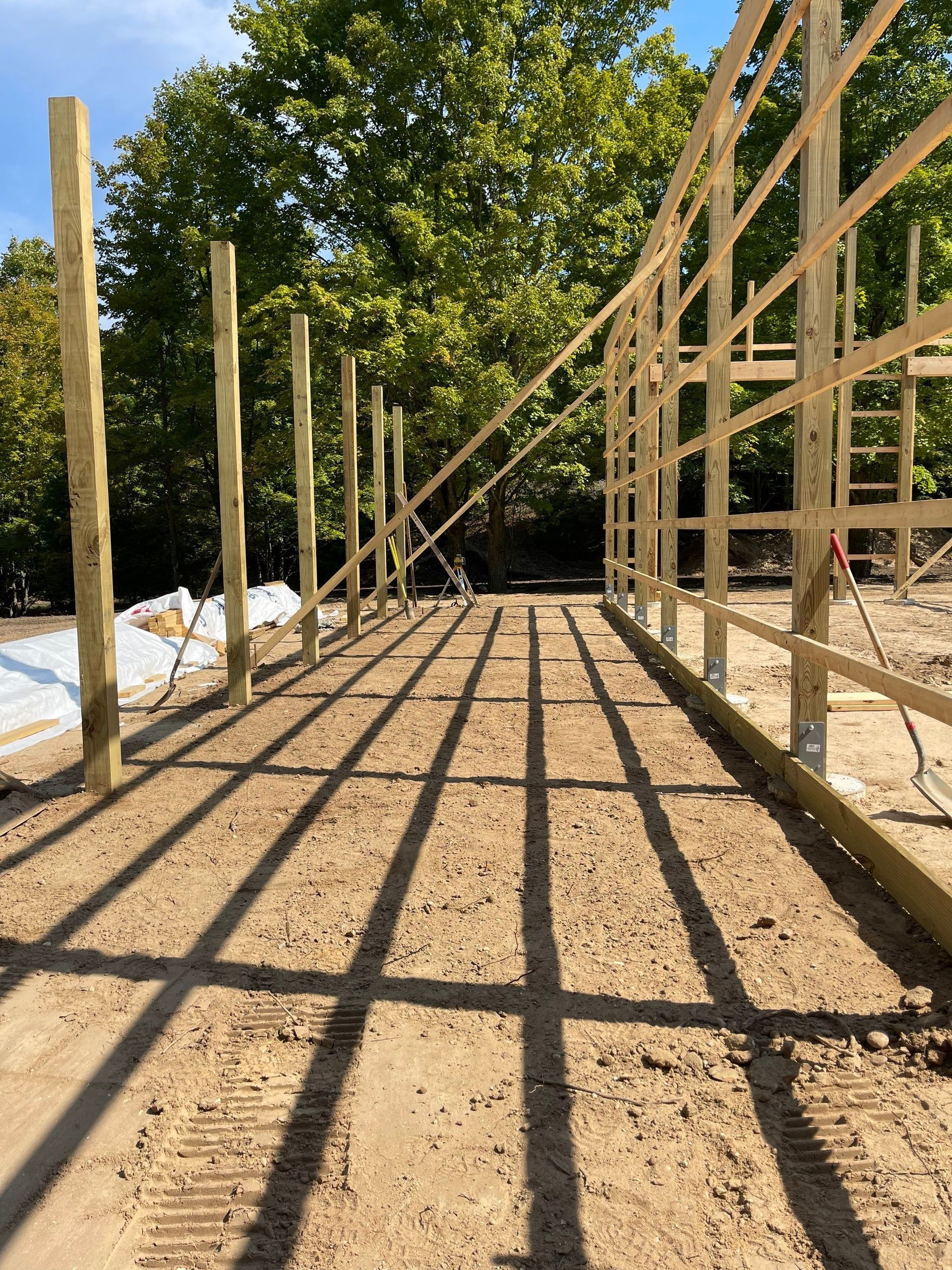 Wooden frame of a building under construction, casting long shadows on the dirt ground, sunny outdoors.